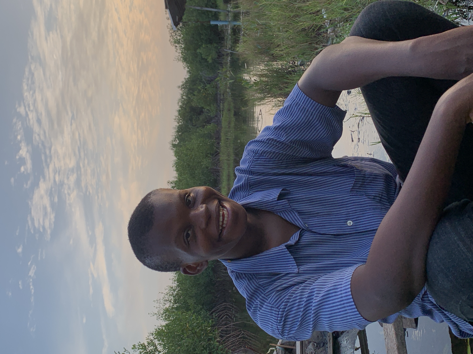 A joyful young male individual is captured in a casual portrait outdoors in Ibasa, Nigeria. He is seated or semi-crouched beside a body of water, looking directly at the camera with a wide, bright smile that shows his teeth. He has a very short haircut and is dressed in a blue and white striped short-sleeved button-up shirt and dark trousers. His arms are bent, resting on his knees.
The setting is a natural, verdant landscape. A calm body of water, likely a river or lagoon, stretches across the midground, with dense green vegetation, including trees and tall grasses, lining its banks. Faint structures or dwellings can be seen in the distance, nestled within the greenery.
The sky, dominating the upper left of the frame (when correctly oriented), displays a soft gradient of colors, transitioning from a pale yellow-orange near the horizon to a light blue, dotted with wispy white clouds. This warm, diffused light suggests the photograph was taken during the golden hour, either in the early morning or late afternoon. The overall mood is serene and natural, enhanced by the subject's happy demeanor. There is no visible text in the image.