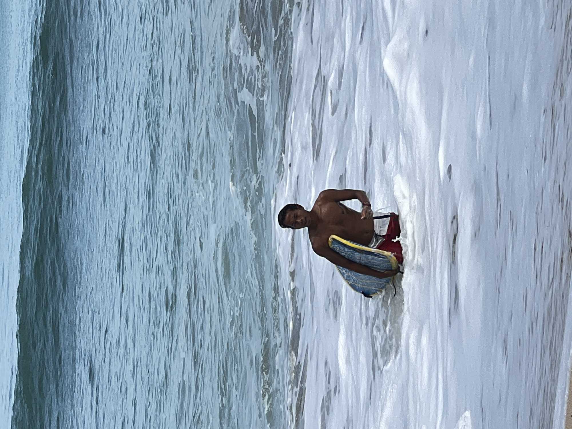 A young, shirtless person with dark hair is standing in foamy water at the beach in Kuta, Indonesia. They are holding a blue and yellow boogie board and wearing red and white swimming trunks. The person is looking towards the right of the frame. The ocean is visible in the background with waves. The lighting suggests it is daytime.