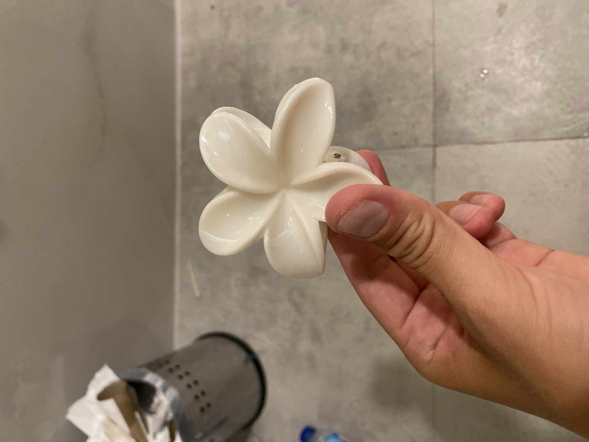 A person's hand is holding a white, five-petal flower-shaped hair clip. The background appears to be a bathroom with tiled flooring and a grey wall. To the left, a stainless steel trash can with holes is partially visible, containing crumpled white paper. The lighting suggests an indoor environment. The scene does not provide any information about the city of Aksu, Turkey.