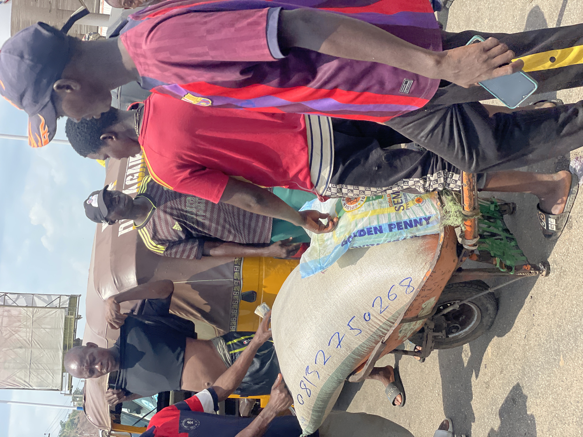 The image shows a group of men gathered around a tricycle loaded with a large sack in Lokoja, Nigeria. The sack, likely containing food, is marked with the words "GOLDEN PENNY" and a phone number. The tricycle is orange and appears worn. The men are dressed in casual attire. Some are looking at the sack, while others appear to be talking or observing. The setting is outdoors on a paved surface, possibly a street or market area. The weather seems bright.