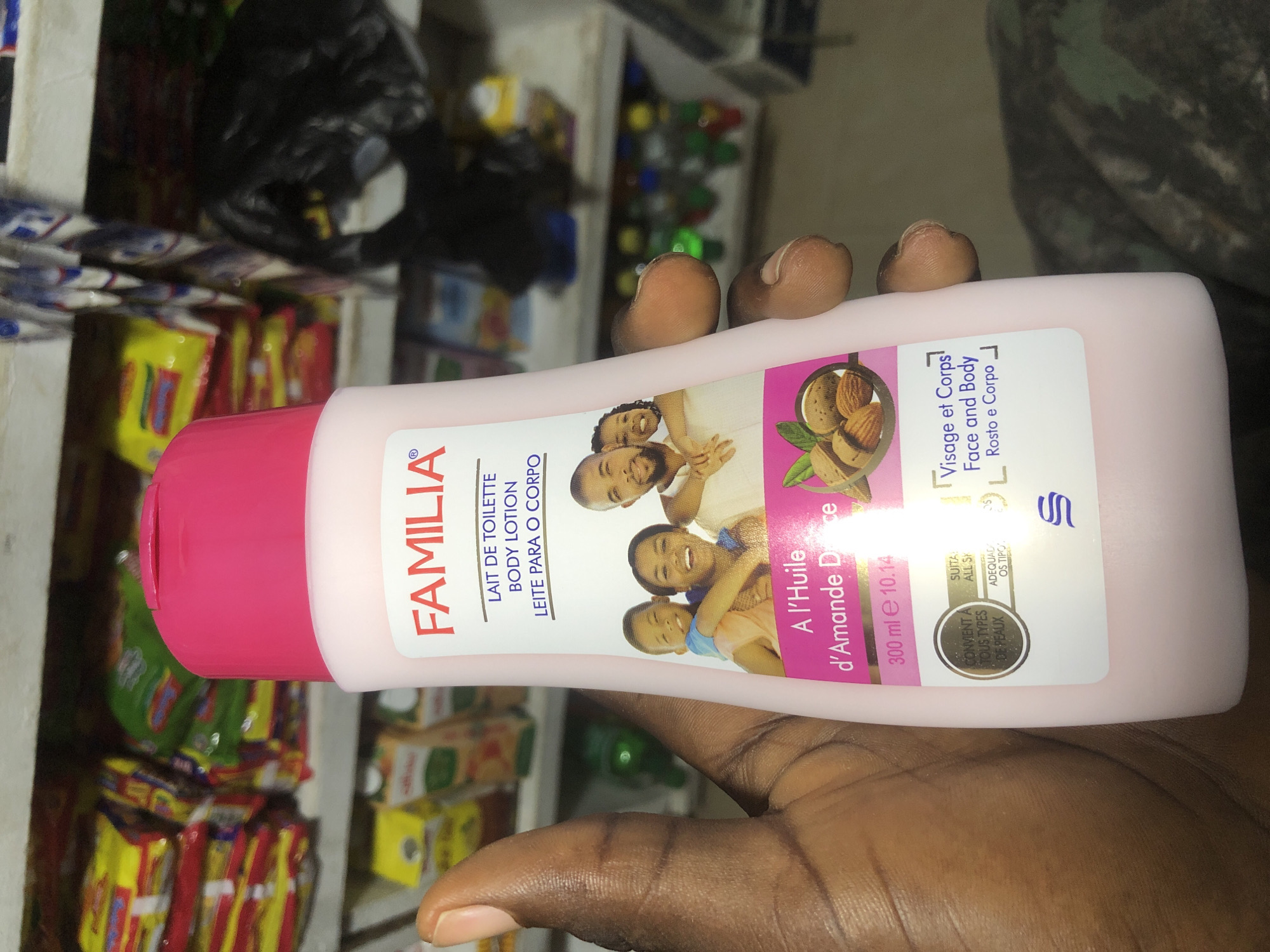 A hand holds a pink bottle of "FAMILIA" body lotion. The bottle has a white label with text and images. The text includes "LAIT DE TOILETTE BODY LOTION LEITE PARA O CORPO". Below that, an image shows a smiling family of four. Further down, there's text about "A l'Huile d'Amande Douce" (Almond Oil) and almonds. The volume is listed as "300 ml e 10.14". There are also small circular icons with text. The background shows shelves stocked with various packaged goods, suggesting a retail environment. The scene appears to be a market or shop.