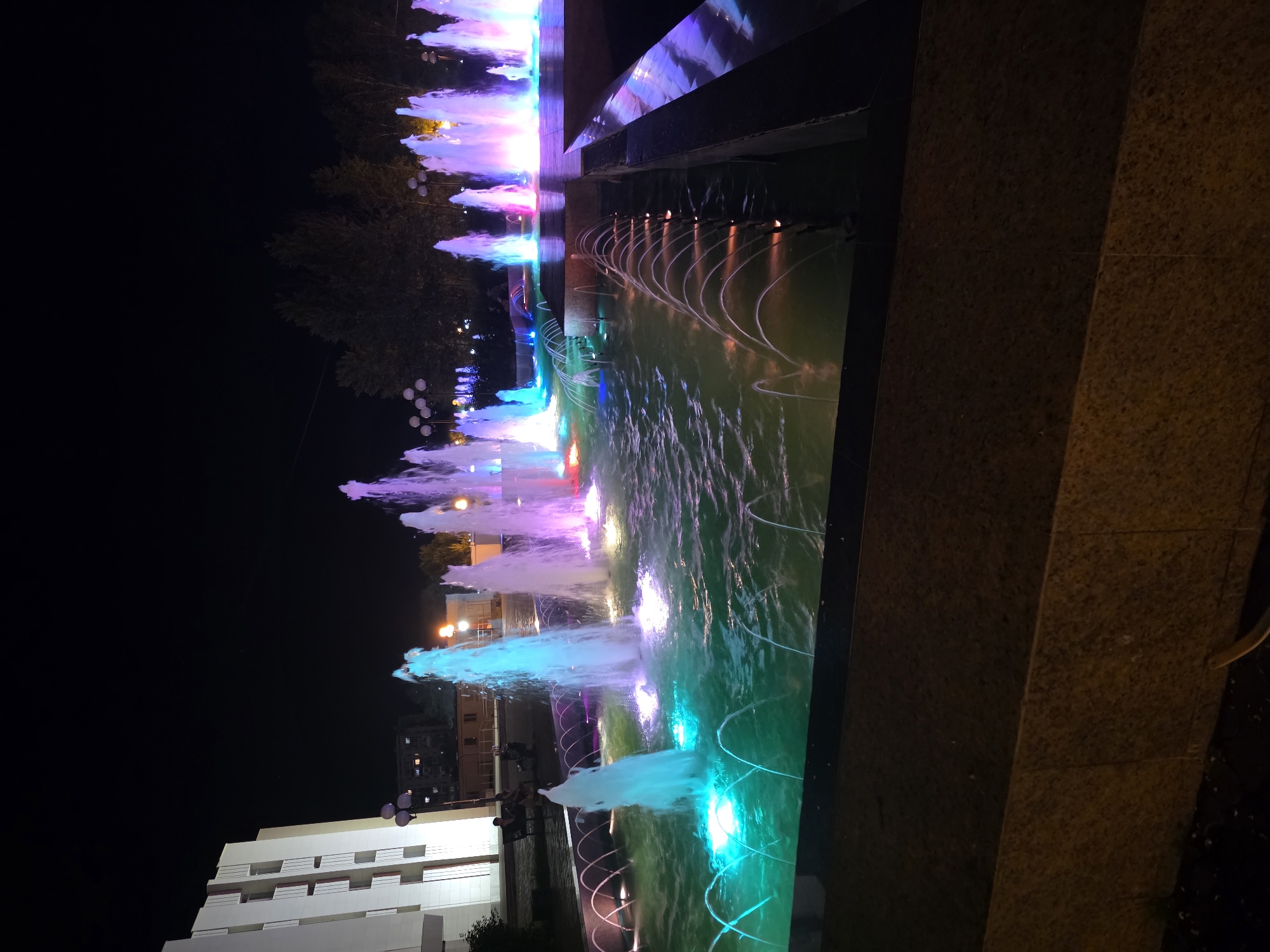 The image depicts a night scene of an illuminated fountain in the city of Komsomolsk-on-Amur, Russia. The fountain features multiple jets of water, lit with vibrant neon colors including blue, pink, and purple. The water in the basin is a striking emerald green. In the background, a modern, white building with horizontal window elements is visible on the left. Several streetlights with spherical lamps are also present. A few people are seated on benches to the left of the fountain, silhouetted against the light. The environment appears to be a public space, possibly a park or a square, with trees visible in the distance. The overall atmosphere is illuminated and colorful, characteristic of a nighttime city attraction.