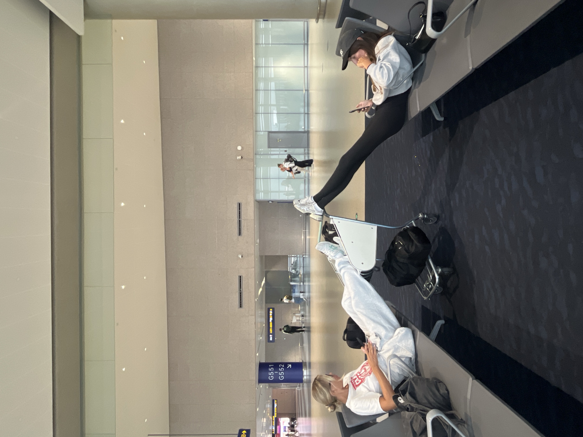 Two young women are seen relaxing in an airport waiting area. The woman on the left, with blonde hair pulled back, wears a white t-shirt and light grey sweatpants. She is looking at her phone and has her legs extended, resting on a luggage cart. The woman on the right, wearing a black baseball cap, a grey hooded sweatshirt, and black leggings, is also looking at her phone with her legs extended. Both are seated on airport benches. The environment is a modern airport terminal with large windows in the background, displaying glimpses of other areas and people. Signs indicating gate numbers G551 and G552 are visible on a blue pillar. Luggage is scattered around them, including a black duffel bag on a silver cart. The floor is covered in a dark, patterned carpet. The lighting suggests it is daytime. This scene is likely at Shanghai Pudong International Airport in Pudong, China.
