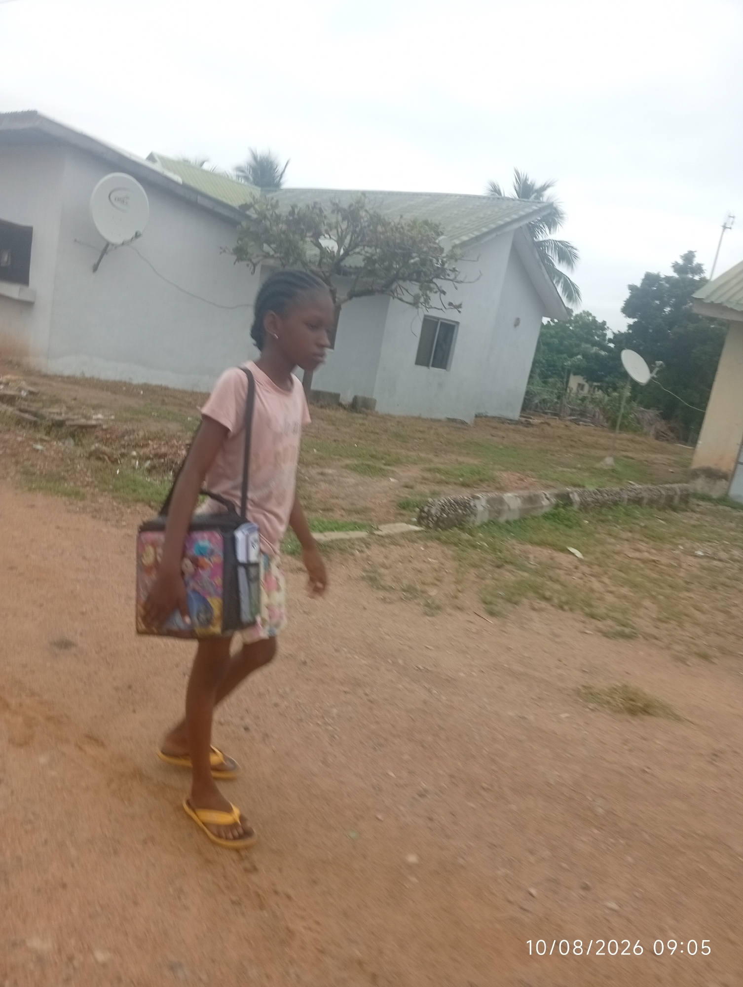 A young girl with braided hair walks on a dirt path, carrying a colorful bag over her shoulder. She is wearing a pink t-shirt and patterned shorts, and yellow flip-flops. In the background, there are houses with satellite dishes, trees, and a cloudy sky. The timestamp "10/08/2026 09:05" is visible in the lower right corner.