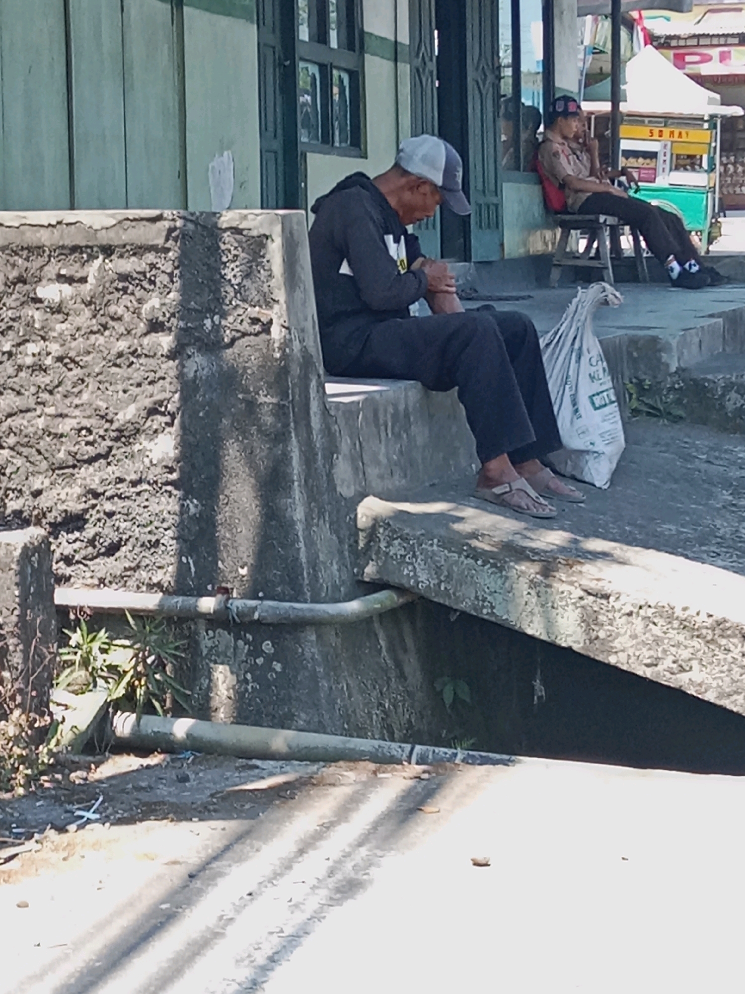 The image depicts a street scene in Bojasari, Indonesia. In the foreground, a man wearing a dark hoodie and a gray baseball cap sits on a concrete ledge, looking down at his hands. He is wearing black pants and light-colored sandals. Beside him, on the ledge, is a large white plastic bag. Further in the background, another individual, possibly a young man, is seated on a wooden stool. He is wearing a light brown uniform with a red backpack and a dark cap. He appears to be looking to his right and may be holding a phone to his ear. Behind him, a food stall with yellow and red signage is visible. The environment appears to be outdoors, with a building and some vegetation in the background. The overall impression is a snapshot of everyday life in a public space.