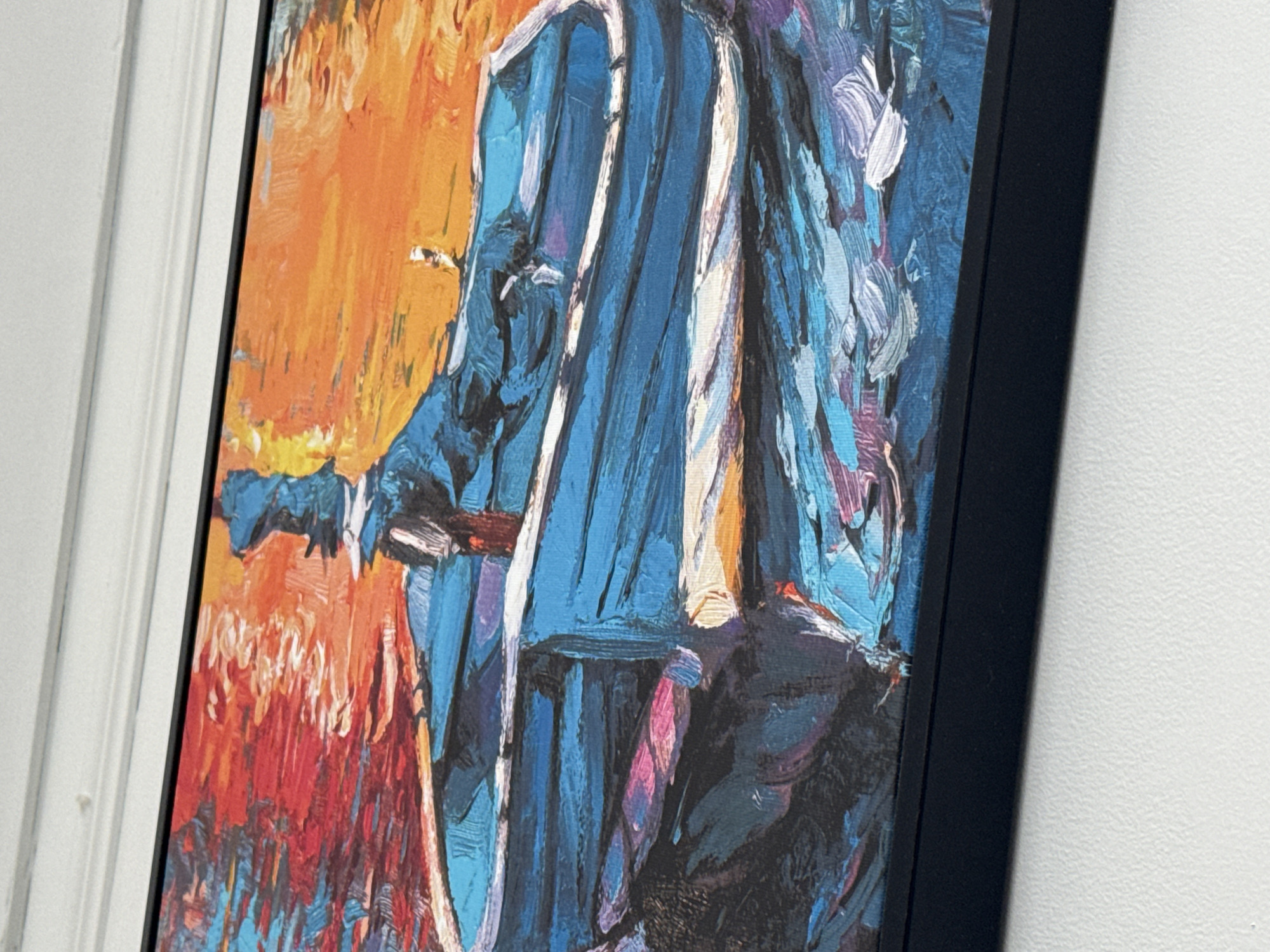 The image shows a close-up, framed oil painting hanging on a white wall. The painting depicts an abstract figure in blues and whites against a backdrop of red and orange hues. The figure's form is suggested rather than explicitly rendered, with thick, visible brushstrokes creating texture and movement. A black frame surrounds the artwork. The location in Đà Nẵng, Vietnam is not discernible from the image.