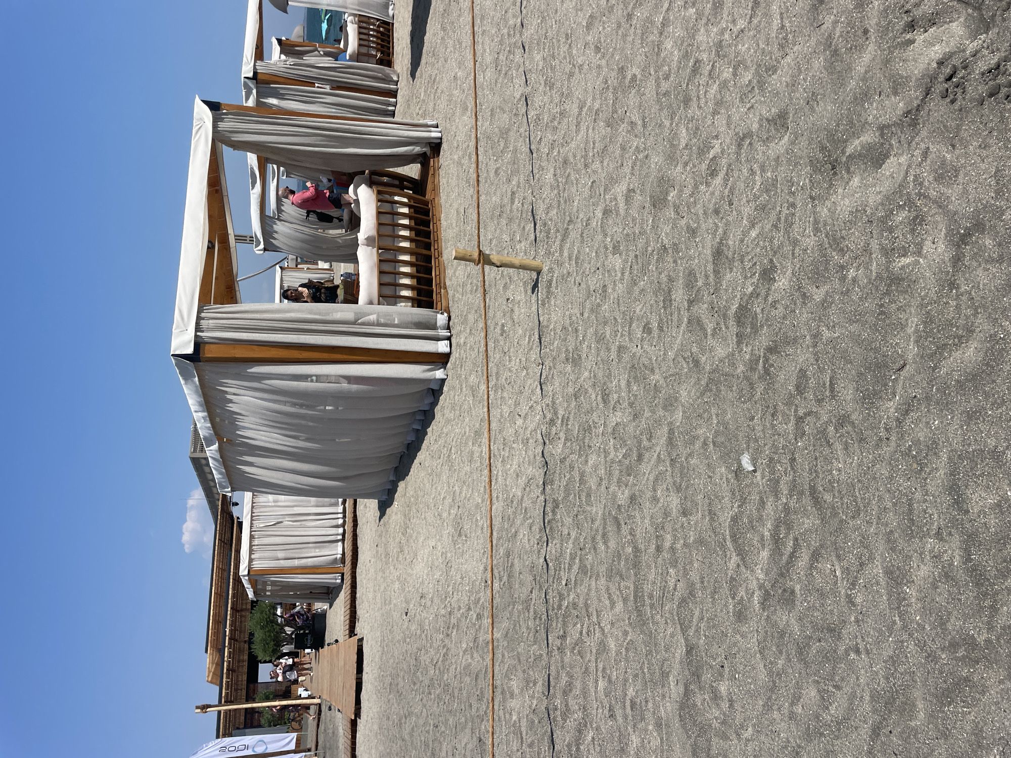 The image depicts a beach resort scene on the coast of Burgas, Bulgaria. In the foreground, a sandy beach is visible with a rope barrier supported by a wooden stake. Several white cabana-style tents with light-colored curtains are set up on the sand, providing shaded seating areas. Inside one tent, a person in a pink shirt and dark shorts is standing, and another person in a dark dress is seated. In the background, a wooden boardwalk leads to a structure with a thatched roof, possibly a bar or restaurant, and people are walking on it. A white flag with blue text is also visible. The sky is clear blue.