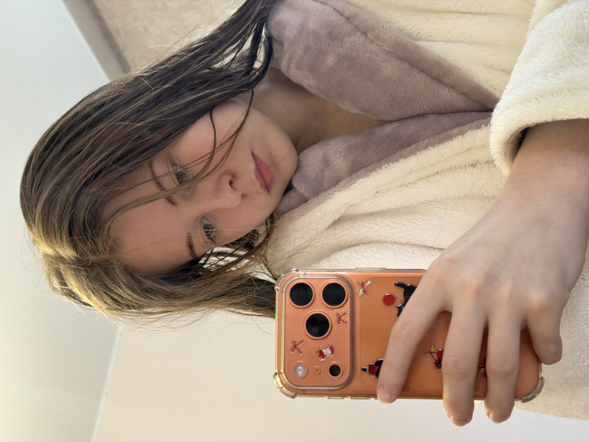 This mirror selfie captures a young individual, likely female, with long, damp brown hair, wearing a cozy cream-colored fluffy bathrobe with a light brown interior. She has light-colored eyes and a soft, slight smile, gazing directly into the camera.
She is holding an orange smartphone in her right hand, which features a clear protective case adorned with several decorative stickers. Notable decals on the case include a prominent red letter "K" near the camera lenses, alongside other small, colorful designs such as red bows and what appears to be a black cat-like figure with red accents. The phone's multi-lens camera system is clearly visible.
The image is taken indoors, against a plain, light-colored background suggesting a wall or ceiling, indicative of a casual home setting, possibly a bathroom or bedroom. The composition is angled, with the subject's head tilted, conveying a relaxed and candid moment, perhaps shortly after a shower or bath. The provided location context is Perm, Russia.