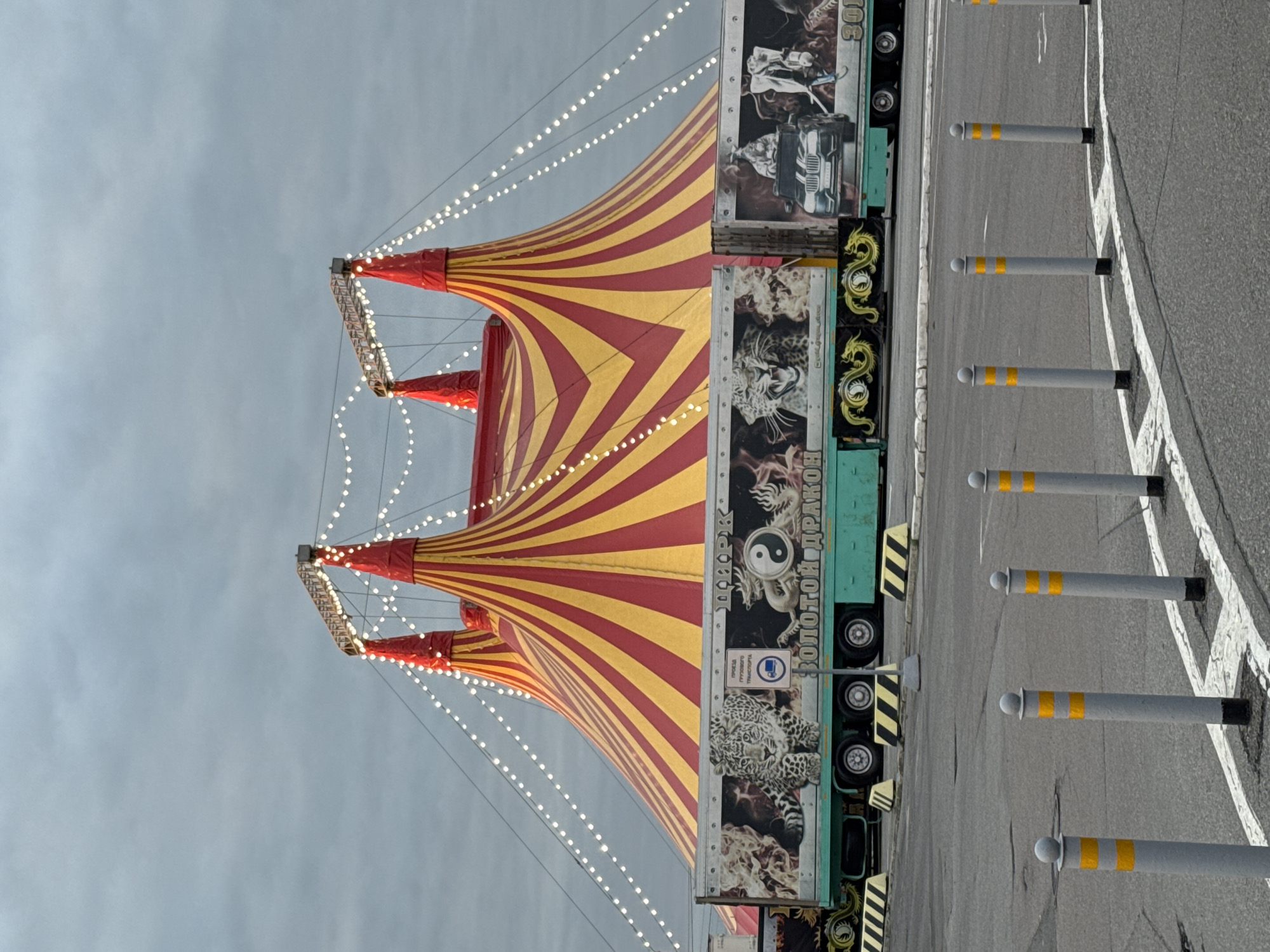 The image, correctly oriented, presents a colorful circus tent set against a dull, overcast grey sky in what is identified as Saint Petersburg, Russia. The dominant subject is a large circus big top, featuring striking red and yellow spiraling stripes that create a sense of movement. Small, white lights are strung along wires extending from the tent's multiple peaks, illuminated despite the daytime appearance, suggesting either an early morning, late afternoon, or a particularly dim day.
Adjacent to the tent are several long, teal-colored trailers or custom vehicles belonging to the circus. These vehicles are elaborately decorated with distinctive artwork, including a fierce leopard or jaguar, intricate golden dragons, and a prominent yin-yang symbol. Visible Cyrillic text on the trailers confirms the presence of a circus: "ЦИРК" (CIRCUS) and "ЗОЛОТОЙ ДРАКОН" (GOLDEN DRAGON), indicating the circus's name. Another visible sign, a blue circle with a truck icon and the text "ПРОЕЗД ГРУЗОВОГО ТРАНСПОРТА," denotes "TRUCK PASSAGE" or "VEHICLE ENTRY" for cargo transport.
A paved asphalt road with white painted lines runs parallel to the trailers, separated by a row of grey bollards with reflective yellow stripes. The scene depicts a temporary outdoor setup for a circus, devoid of people, with the focus on the vibrant tent and detailed transport vehicles under a muted, possibly drizzly, sky.