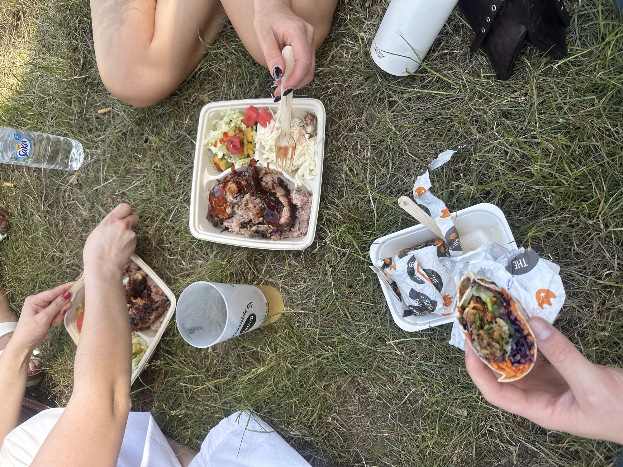 The image depicts an outdoor scene in London, United Kingdom, where people are having a picnic on the grass. Visible are two individuals seated on the grass and another standing while holding a wrap. Three takeaway containers are present, containing food items like jerk chicken, rice, salad, and wraps. There are also a plastic bottle of water, a cup, and a white metal water bottle. The atmosphere suggests a casual outdoor meal.