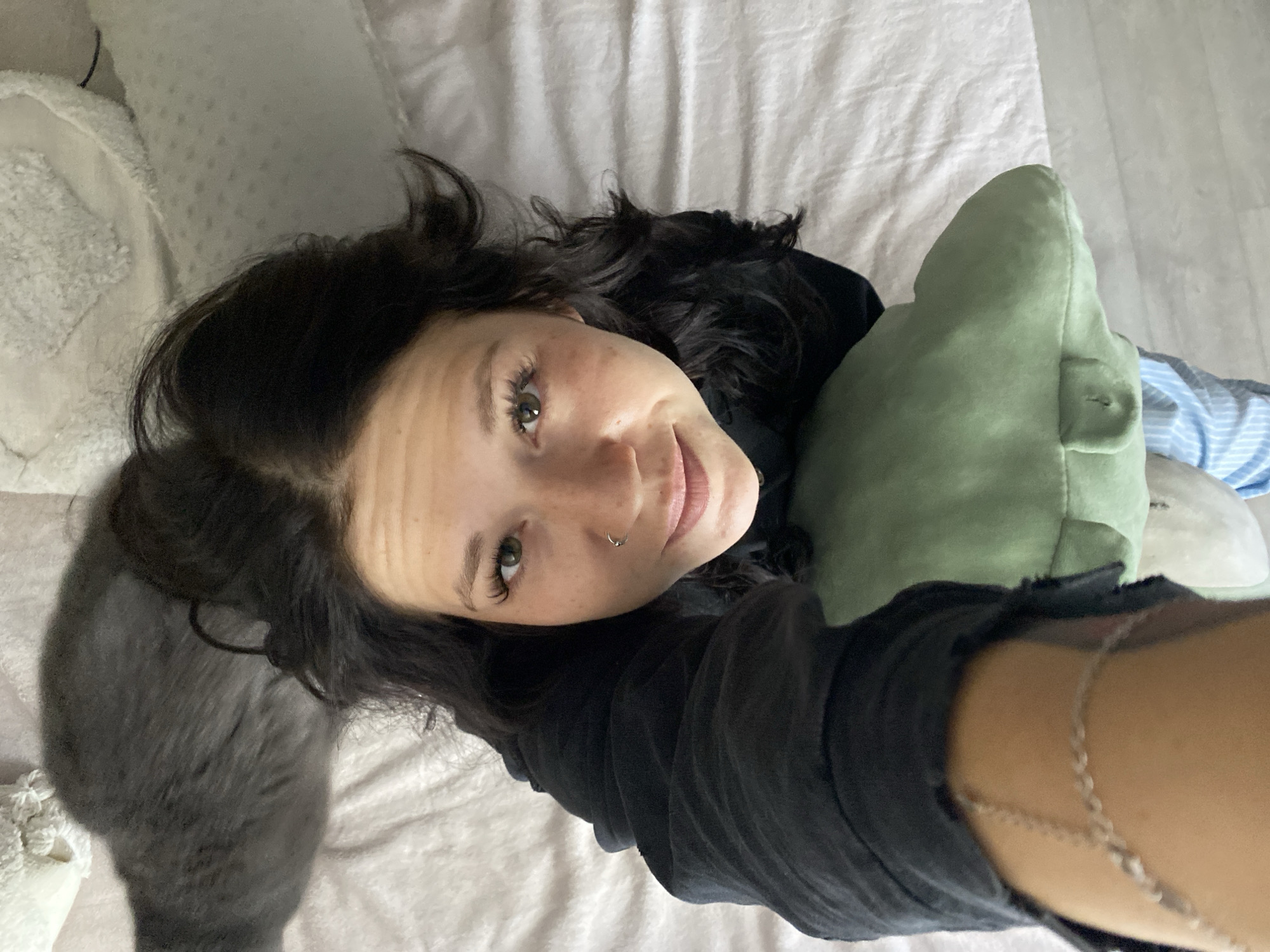 A high-angle, close-up shot captures a young woman lying on a white, rumpled sheet, with her head tilted back and her face looking towards the camera. She has dark, wavy hair, green eyes, and light skin with freckles. She is wearing a black hoodie and a silver chain around her wrist. A grey cat is visible behind her head. A green pillow is positioned in front of her. The background consists of bedding and a hint of a light-colored, textured surface. The scene appears to be indoors, possibly a bedroom. There is no discernible indication of the city of Chaikovsky, Russia.