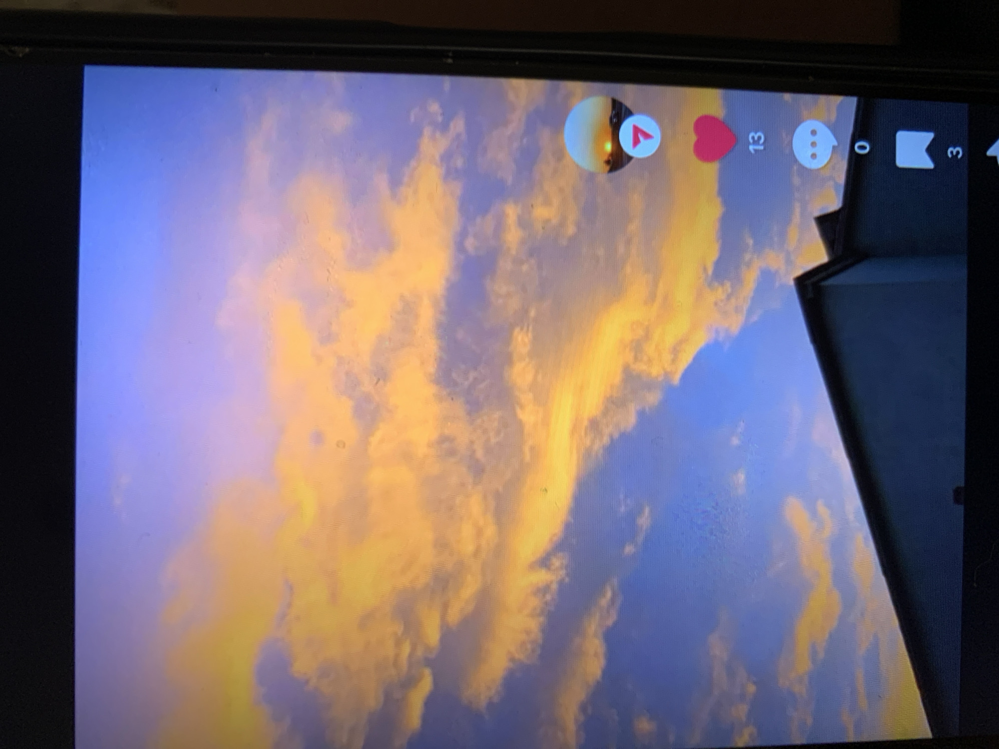 The image displays a cell phone screen showing a social media interface, likely TikTok, with a sunset scene in the background. The sky is filled with dramatic clouds illuminated by the setting sun, casting hues of orange and yellow against a blue sky. In the foreground, the dark silhouette of a building's roofline is visible. On the right side of the screen, social media interaction icons are displayed: a red heart with the number "13" below it, a speech bubble icon with the number "0" below it, and a bookmark icon with the number "3" below it. There is also a circular icon showing a sunset with a red play button. The image does not contain any information or visual cues that would allow for the identification of the city of Lymanka, Ukraine, or any specific location within it.