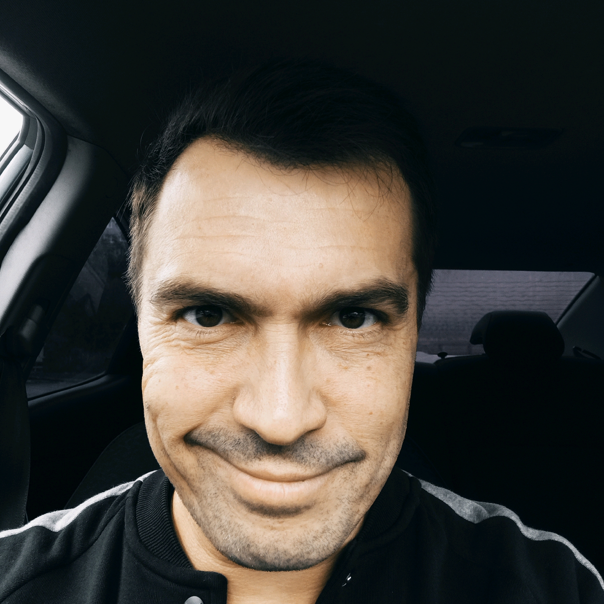 A mid-shot, eye-level selfie taken from inside a car. The subject, a man with dark hair and brown eyes, faces the camera directly with a slight, closed-mouth smile. He wears a black jacket with gray stripes on the shoulders. The interior of the car is visible, including a dark headliner and part of the passenger seat. The background outside the car window is blurred, suggesting a natural or urban environment, possibly the city of Nizhny Novgorod, Russia, given the context. The lighting is overcast, creating a soft, diffused light on the subject's face.