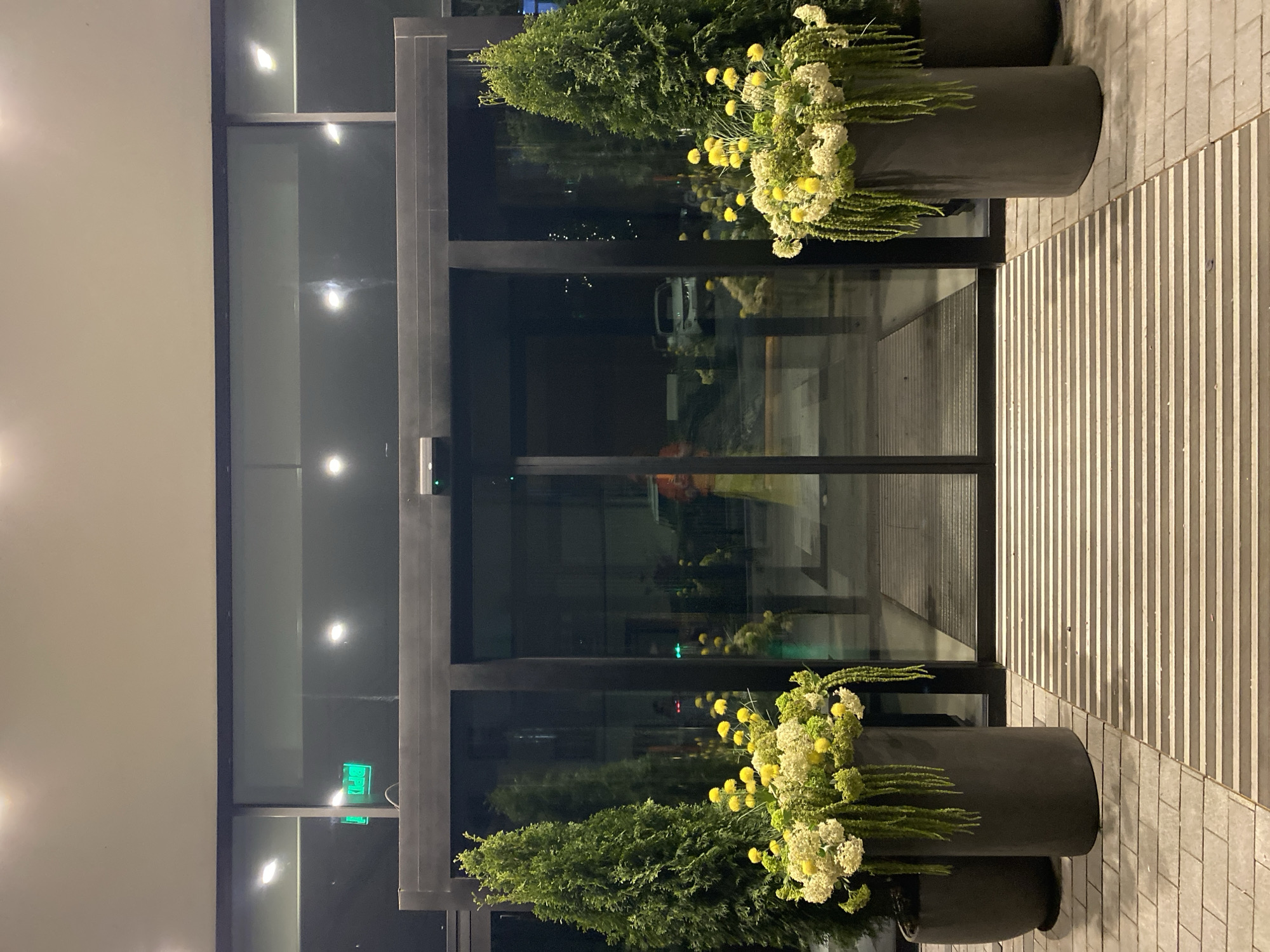An exterior shot of a building at night, potentially in Yekaterinburg, Russia. The entryway features a revolving door with black framing, flanked by large cylindrical planters filled with green shrubs and yellow and white flowers. The building's facade is illuminated, and the walkway leading to the entrance is paved with striped concrete. Reflections in the glass suggest the presence of vehicles and other lights outside.
