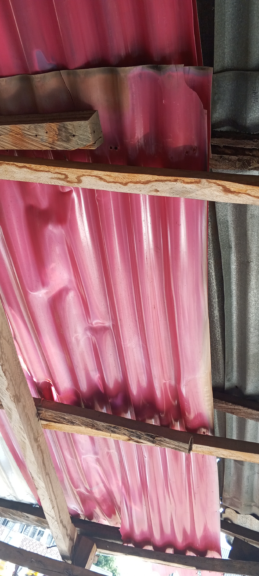 The image shows a close-up, low-angle view of a damaged, corrugated pink plastic roof supported by weathered wooden beams. The plastic sheeting is creased, torn in places, and appears to be partially melted or discolored along the top edge, suggesting exposure to heat. To the right of the pink roof, a section of corrugated galvanized metal roofing is visible. The scene is likely from a structure in Jalingo, Nigeria, possibly indicating damage from an event like a fire, given the discolored and melted appearance of the plastic.