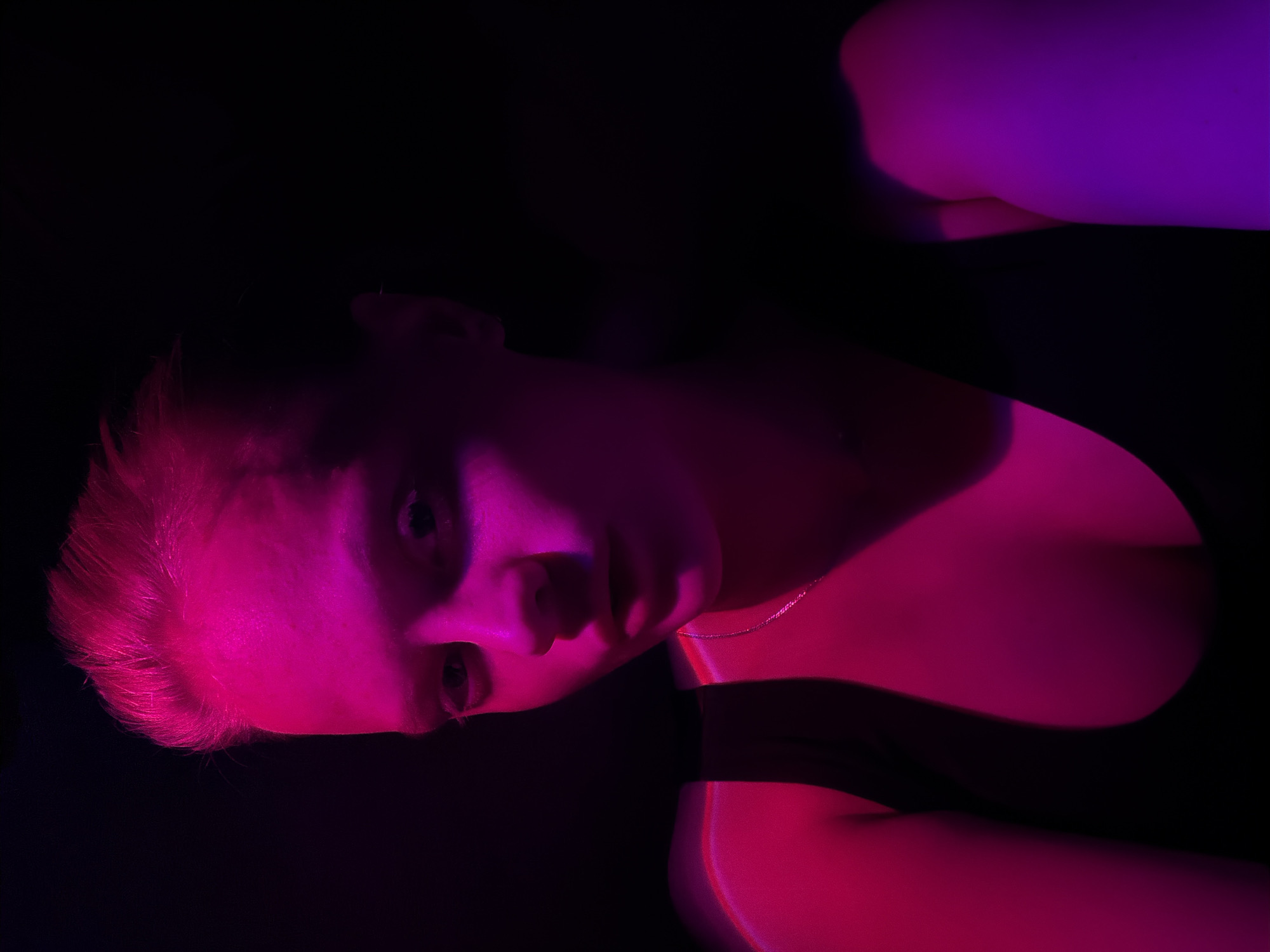 A person with light skin and short, light hair is illuminated by pink and purple light against a dark background. The person is angled, with their face and part of their chest visible. They appear to be wearing a dark top. The location is not discernible from the image.