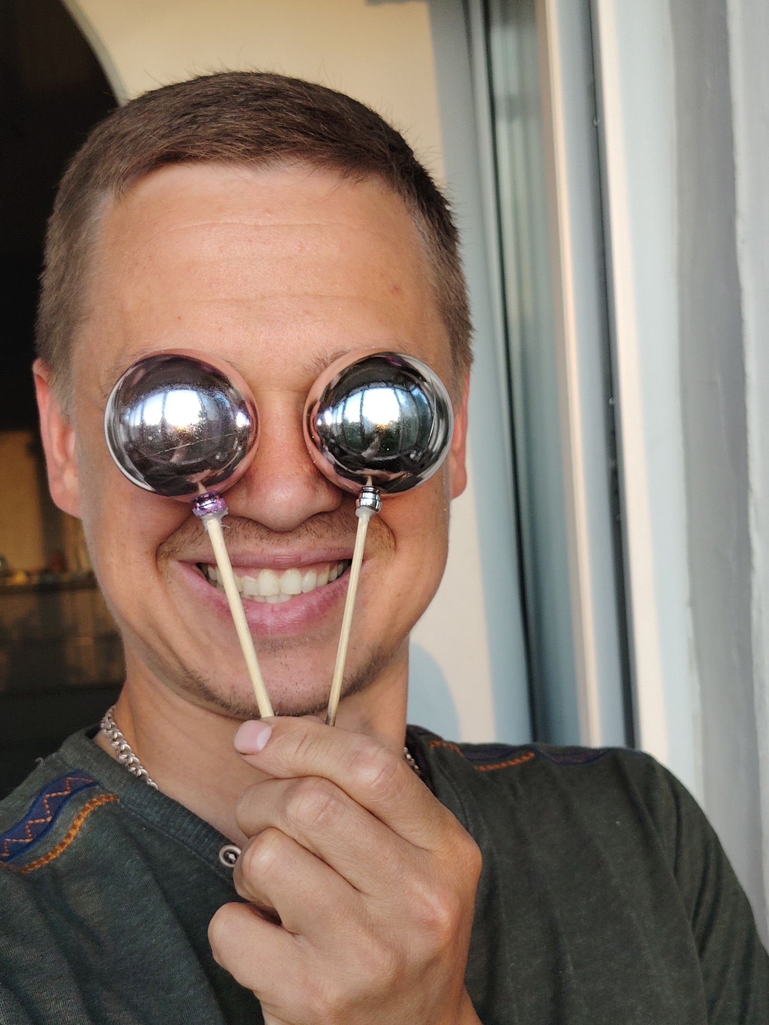 A man is captured in a close-up, chest-up portrait, playfully holding two shiny, spherical, metallic ornaments on wooden sticks over his eyes. He is smiling broadly, revealing his white teeth, and appears cheerful and amused. His short, brown hair is visible, along with light stubble on his face.
He is wearing a dark green, short-sleeved t-shirt featuring subtle embroidered patterns in orange and blue on the left shoulder, and a silver chain necklace around his neck. His hand, showing knuckles and fingers, holds the wooden sticks just below the reflective spheres. The spheres themselves gleam, reflecting ambient light and hints of the surrounding environment, including what appears to be a window.
The background is softly blurred but suggests an indoor or semi-outdoor setting, possibly a balcony, with natural light streaming in, indicating it is daytime. A light-colored window or door frame is visible on the right side of the frame, contrasting with darker, indistinct areas on the left that suggest an interior space. The overall impression is one of lightheartedness and joy. The scene is set in Chaikovsky, Russia. No visible text is present in the image.