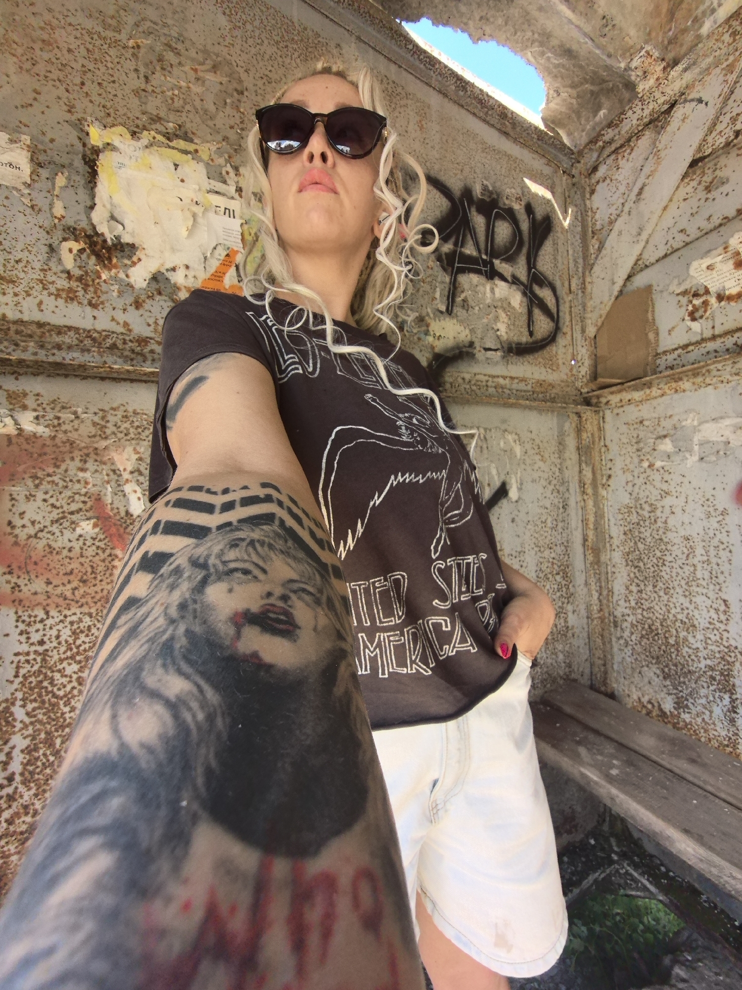 The image depicts a young Caucasian woman with blonde curly hair, wearing black sunglasses, a dark t-shirt with a graphic print, and white denim shorts. She is standing in what appears to be a derelict, open-air structure with rusty metal walls and a hole in the ceiling exposing blue sky. Graffiti is visible on the walls. The woman's left arm is prominently tattooed with a black and grey portrait of Marilyn Monroe. The setting suggests an urban environment, possibly an abandoned building or a disused structure. There are no other individuals present in the image. The overall impression is one of urban decay and personal expression through fashion and tattoos.