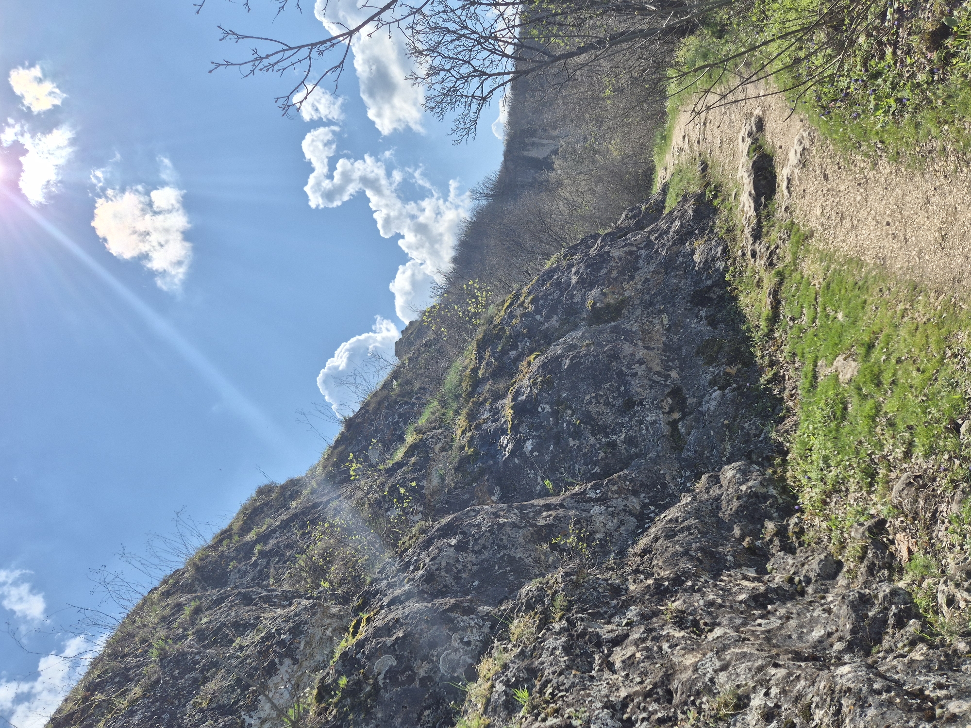 The image captures a rugged, rocky terrain under a bright, partly cloudy sky, with a prominent cliff face covered in moss and sparse vegetation. A dirt path runs along the top of the cliff, bordered by grass and foliage. Above the cliff edge, a forested area can be seen. The sun is visible in the top left corner, casting rays across the sky. Based on the presence of rock formations and the description, it is likely taken at the Rock-hewn Churches of Ivanovo, a UNESCO World Heritage Site near Ivanovo, Bulgaria.