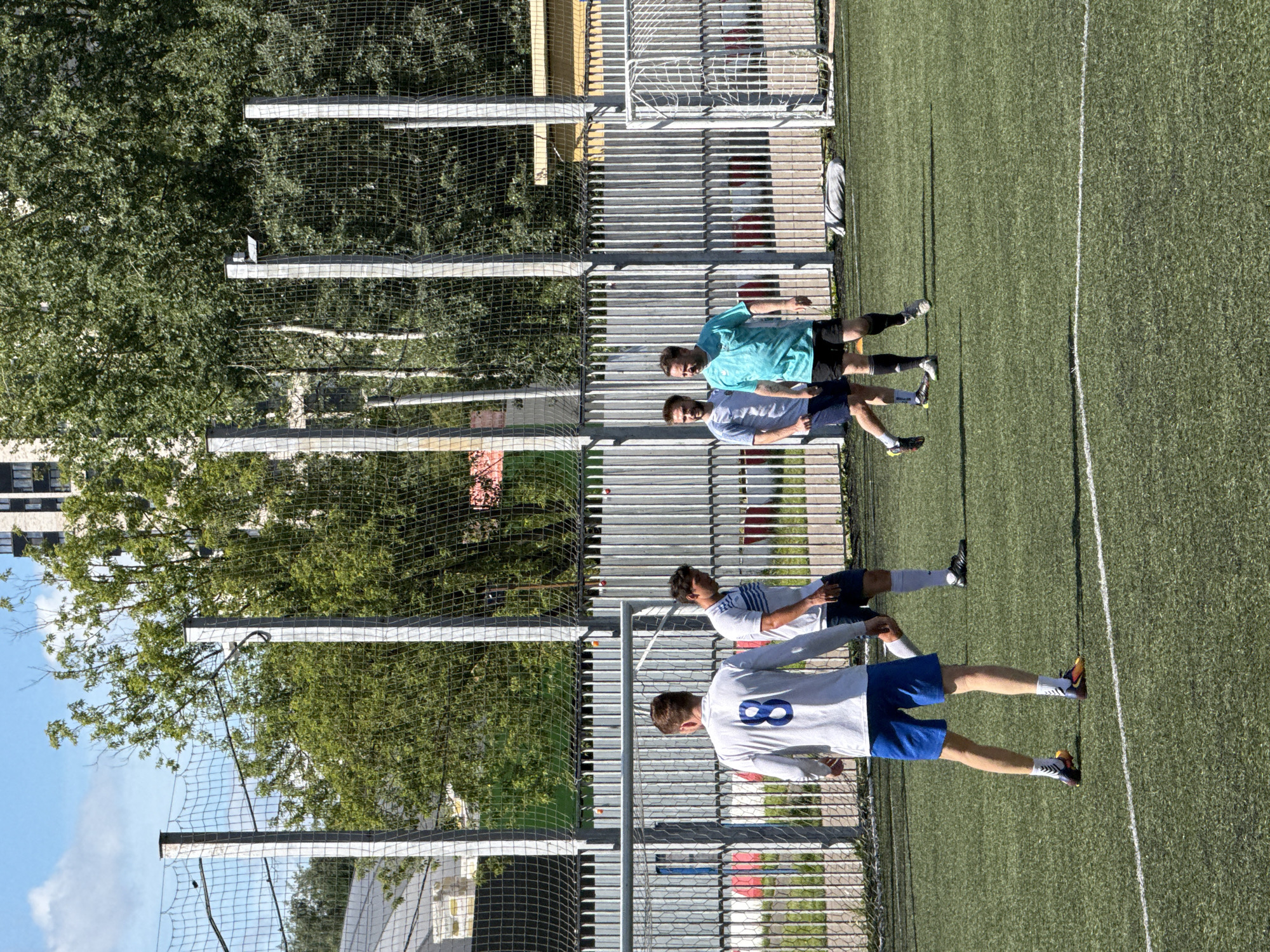 Four men are playing soccer on a synthetic turf field, with a chain-link fence and trees in the background. The man on the far left is wearing a white long-sleeved shirt with a blue number '8' on the back, blue shorts, white socks, and multi-colored cleats. The man next to him is wearing a white t-shirt with blue stripes, dark shorts, and white socks. The third man from the left is wearing a light gray t-shirt and dark shorts. The man on the far right is wearing a turquoise t-shirt, black shorts, and black socks. The field has white markings. In the background, there is a white chain-link fence, trees, and a tall building. The sky is blue with some clouds.