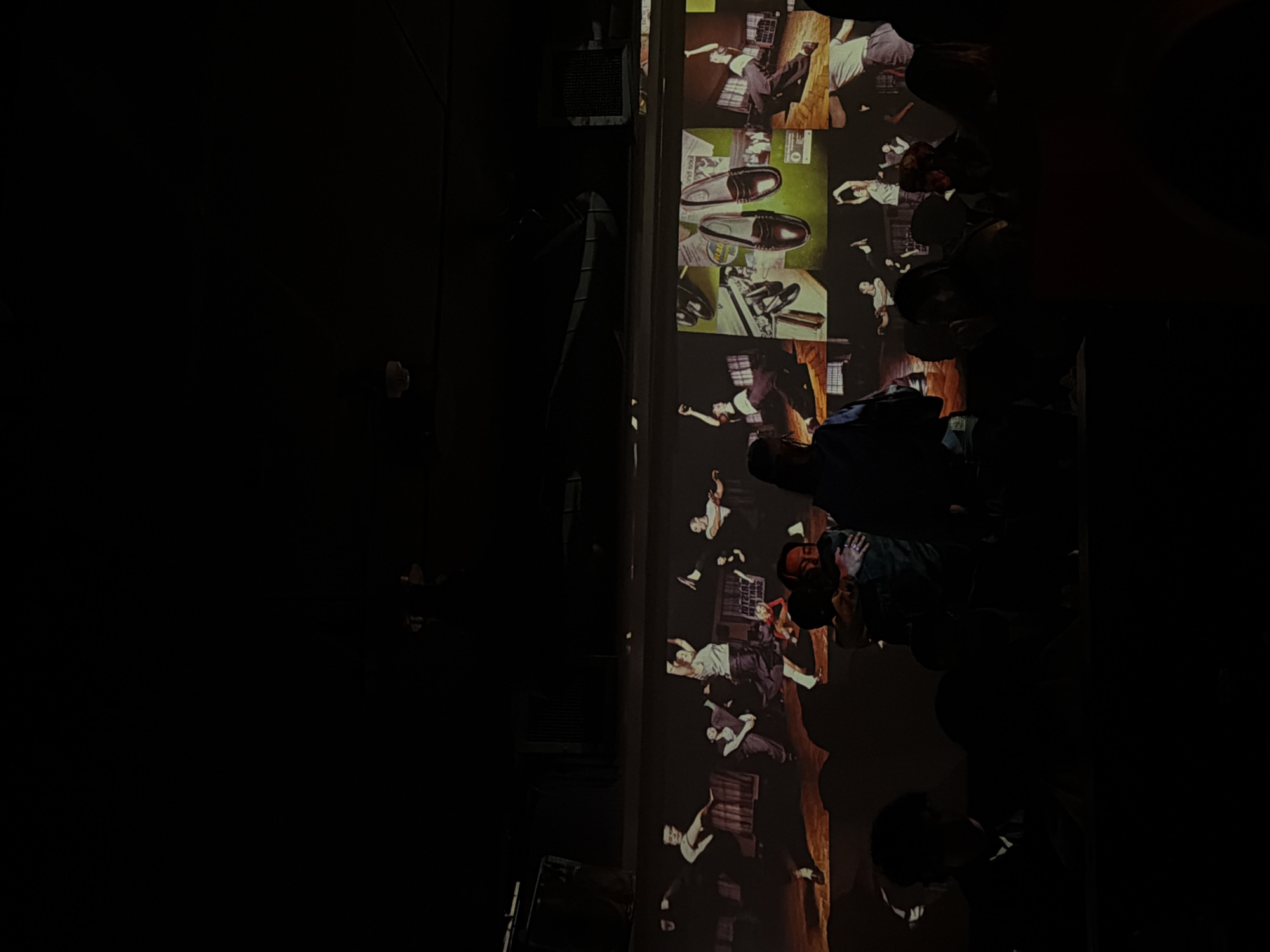 A low-light, indoor scene depicts a group of people watching a projected display. The projection features images reminiscent of a stylized fashion shoot or advertisement, presenting figures in dynamic poses and articles of clothing such as shoes, juxtaposed with what appear to be snapshots of urban settings or interiors. The audience members are partially visible in the dark foreground and are interspersed among the projected images. The locale is assumed to be in the city of London, United Kingdom, though this is unconfirmed by visual details alone.