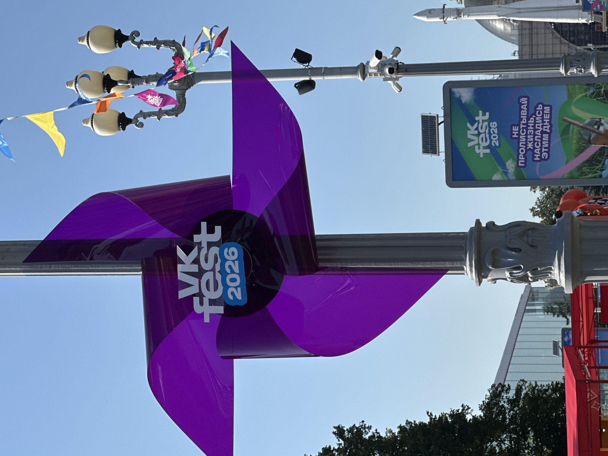 The image shows a bright, sunny day at an outdoor event in Moscow, Russia. In the foreground, a large purple pinwheel decorated with the "VK fest 2026" logo is attached to a lamppost. The pinwheel has four large, curved blades that twist upwards. Behind the pinwheel, another lamppost is visible, adorned with multiple strings of colorful flags and three ornate light fixtures. Further back, a large digital billboard displays the "VK fest 2026" logo and Russian text. Several security cameras are mounted on the lamppost. To the right, a white rocket sculpture stands tall against the clear blue sky, and a large dome-shaped building is partially visible. In the background, trees and a modern building with a glass facade can be seen. Red tents, possibly for vendors, are set up on the left side of the frame. The overall scene suggests a festive and public gathering.