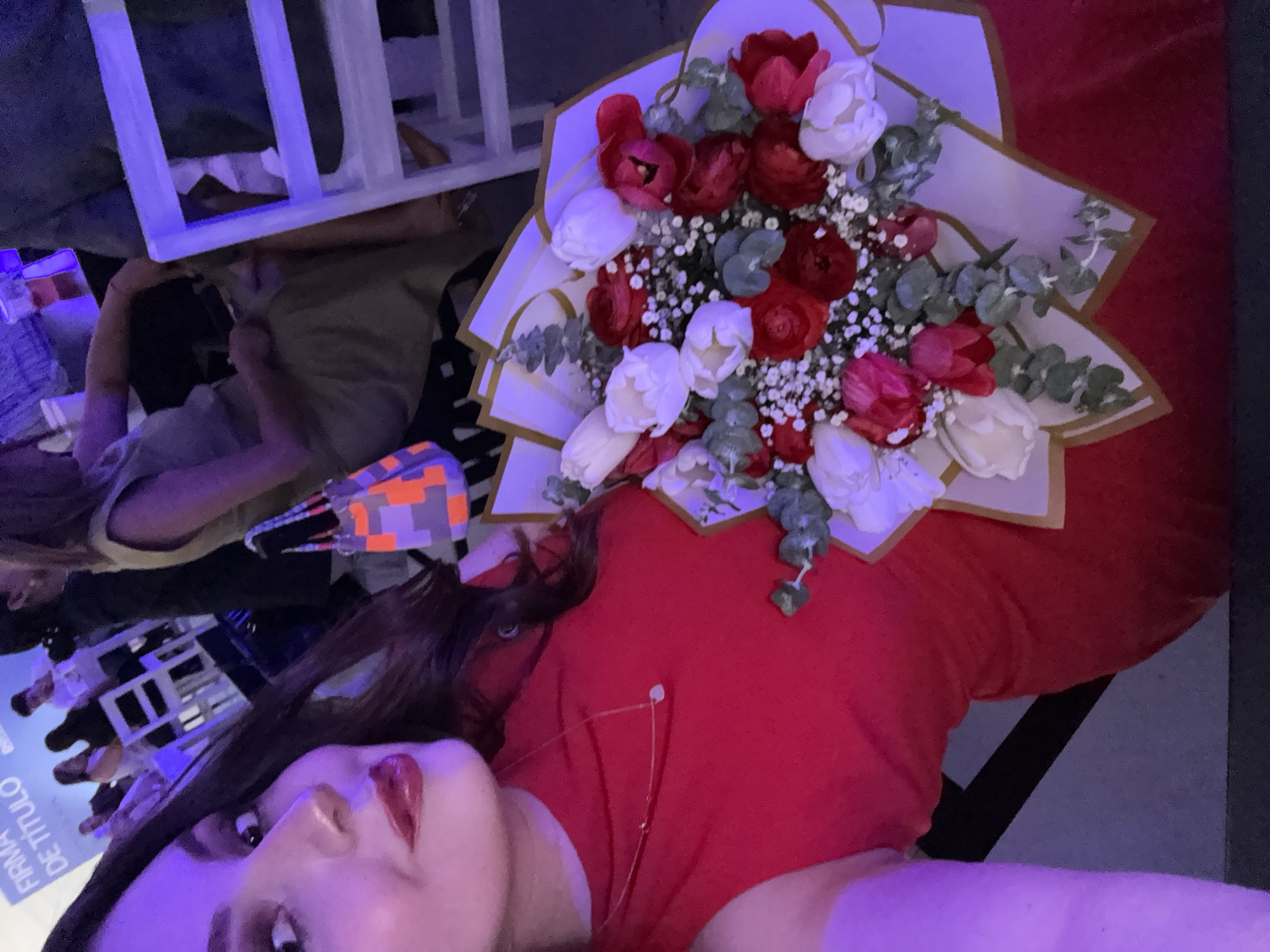 A woman with long dark hair and red lipstick is lying down, wearing a red top. A bouquet of red and white tulips, wrapped in white and gold paper, rests on her chest. The background includes a white wooden structure, a glimpse of another person, a face mask with an orange and gray pattern, and a sign in Spanish, "FIRMA DE TITULO". The location is likely in Mexico City, Mexico.
