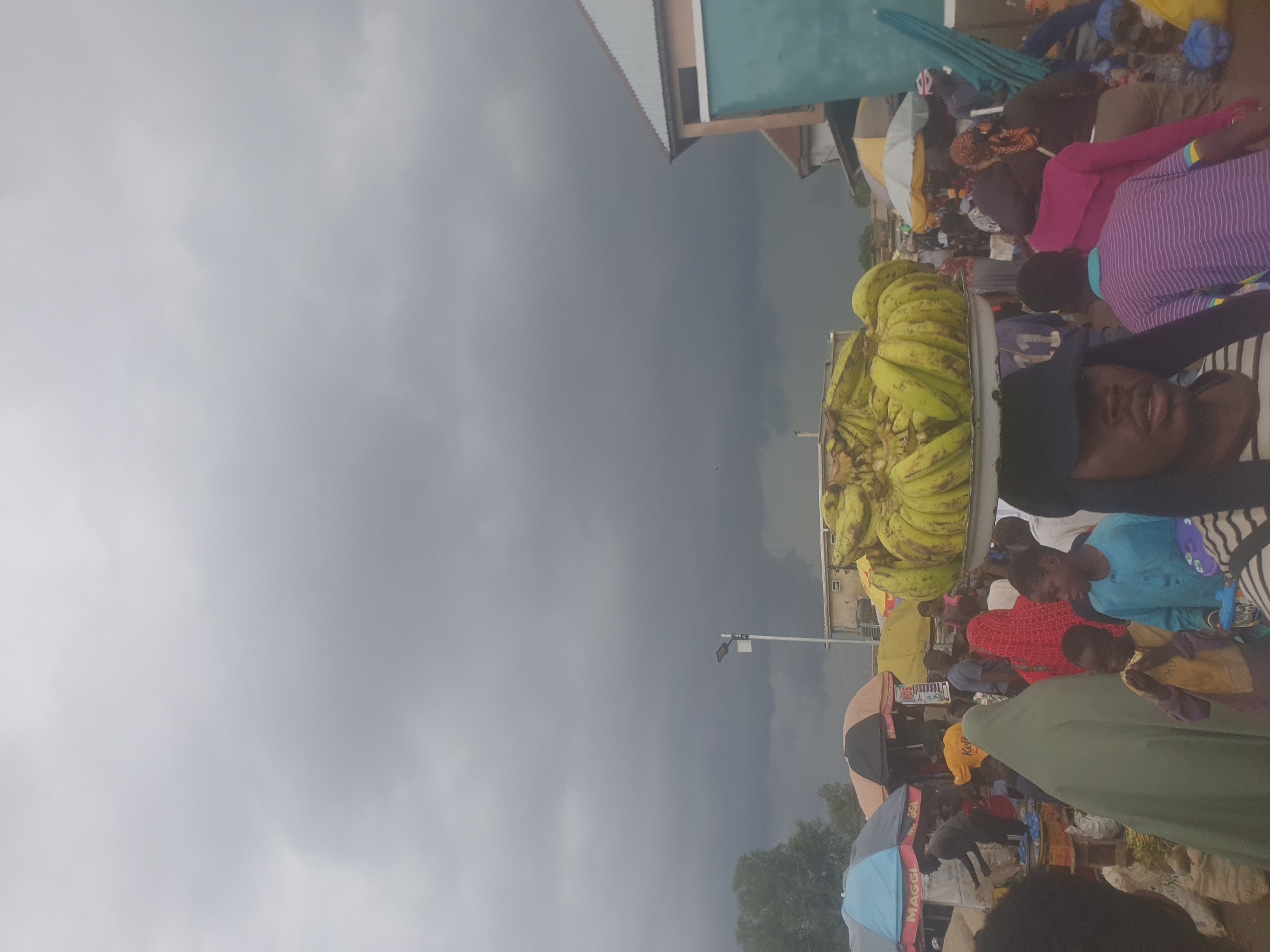This media file depicts an outdoor market scene in Rafin Par, Nigeria. The sky is overcast, indicating cloudy or potentially rainy weather. A prominent display of bananas is visible, suggesting a food market. Several people are present, some appearing to be vendors and others customers. They are gathered around stalls and displays, engaged in activities typical of a market environment. Clothing colors are varied, with a woman in a pink top and another in a purple striped top being noticeable. The overall atmosphere suggests a busy marketplace. There is some visible text on what appears to be merchandise or signage, but it is not clearly legible. The scene seems to be taking place during daylight hours, although the overcast sky makes it difficult to determine the exact time of day. The presence of umbrellas and awnings suggests a need for shelter from the sun or rain.