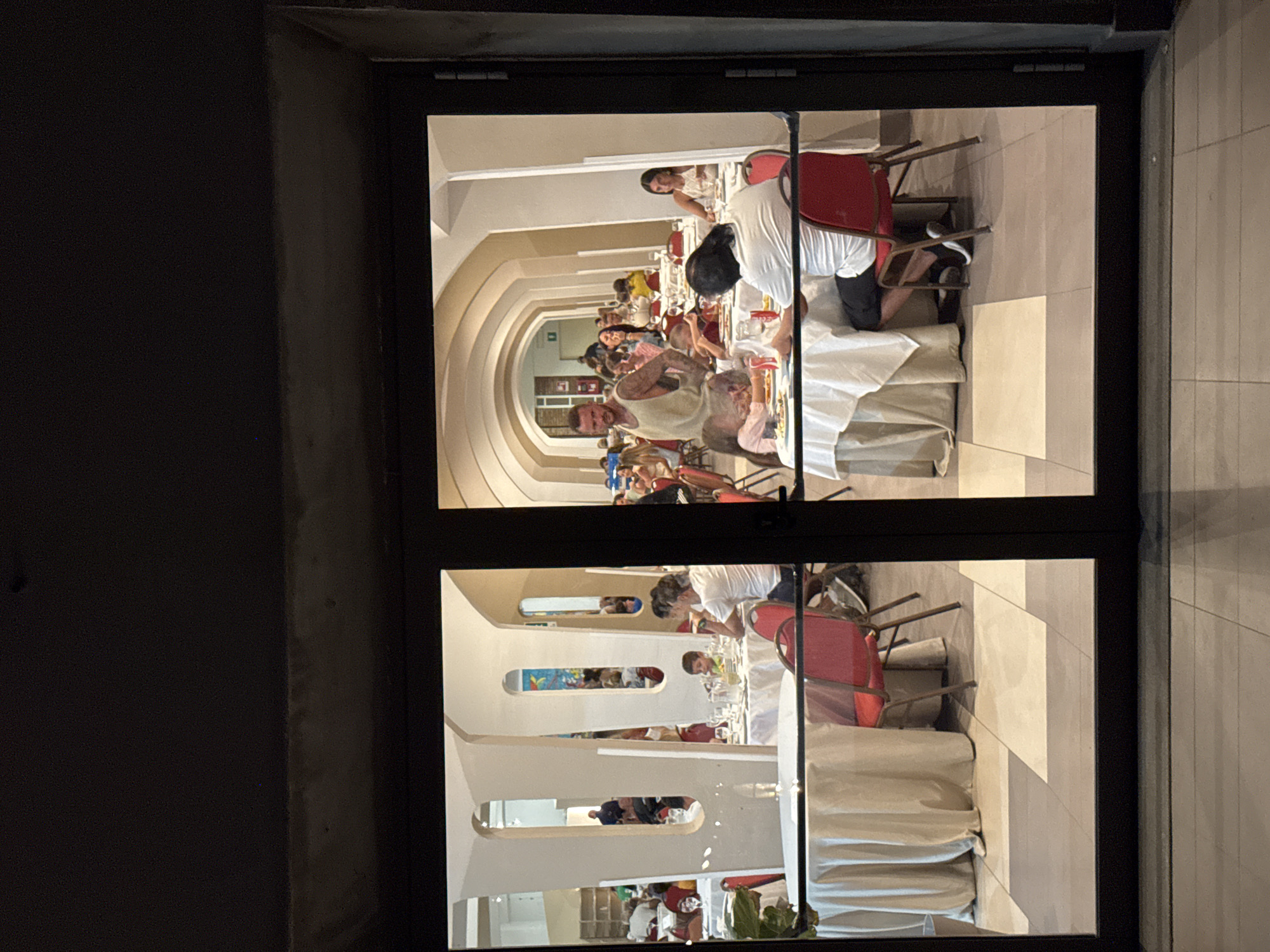 A group of people are dining at tables covered with white tablecloths in a brightly lit, arcaded hall that appears to be a restaurant or banquet hall. The hall has white columns and arched ceilings. In the foreground, a table is set with plates, glasses, and what appears to be food. A person in a white shirt and black shorts is seated at this table. To the left, another table is visible with red chairs. In the midground, a man with tattoos on his arms and wearing a light-colored tank top stands, possibly serving or observing. Several other diners, including children, are seated at tables further back. The scene is viewed through a glass door, with the dark frame of the door visible in the foreground and background. The flooring is tiled in a checkered pattern of light and dark squares. This scene is located within the restaurant of the Costa Blu Hotel in Capaci, Italy.