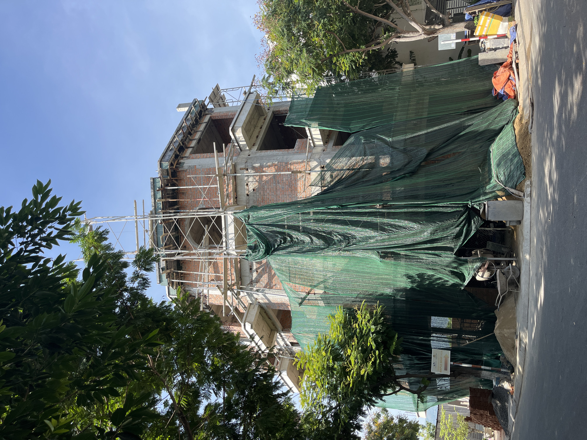 The image shows a construction site in Nam Nha Trang Ward, Vietnam, with a multi-story building under construction. The building is partially covered with green construction netting, obscuring some of the brickwork and scaffolding. Several trees with green foliage are visible in the foreground and around the site. Piles of sand and bricks are present near the building, along with a cement mixer and a wheelbarrow. The sky is clear and blue.