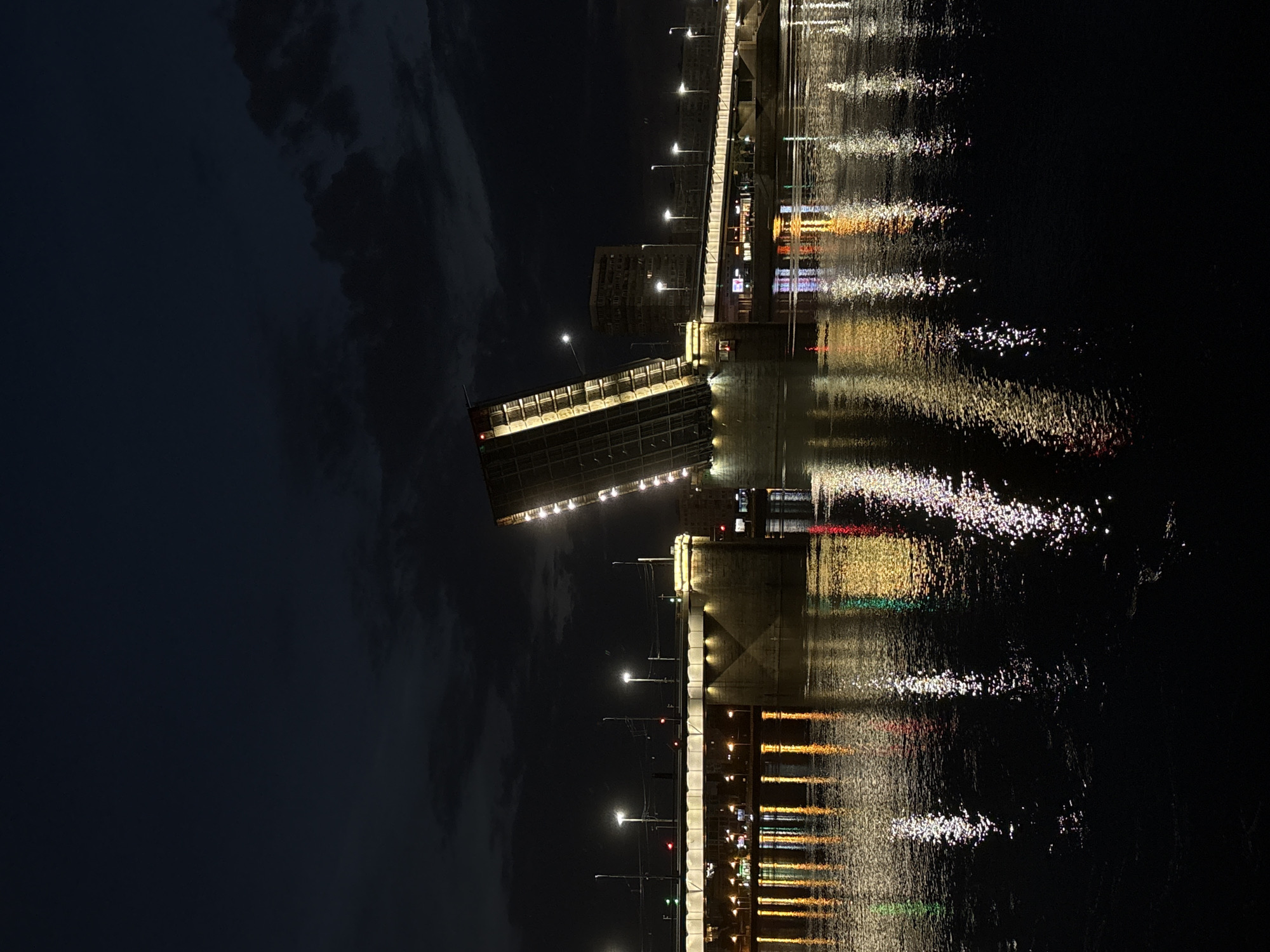 The image depicts a nighttime scene in the city of Otradnoye, Russia, focused on a bascule bridge, also known as a drawbridge, spanning a body of water. The bridge is in the process of being raised, with one section significantly tilted upwards, revealing its mechanical structure illuminated by numerous lights. The water's surface reflects the lights from the bridge and surrounding buildings, creating a shimmering, colorful spectacle. A few distant buildings and streetlights are visible on the opposite bank, under a dark, cloudy sky. There are no people visible in the image. The overall impression is one of urban nighttime ambiance, highlighting the engineering and visual appeal of the raised bridge.