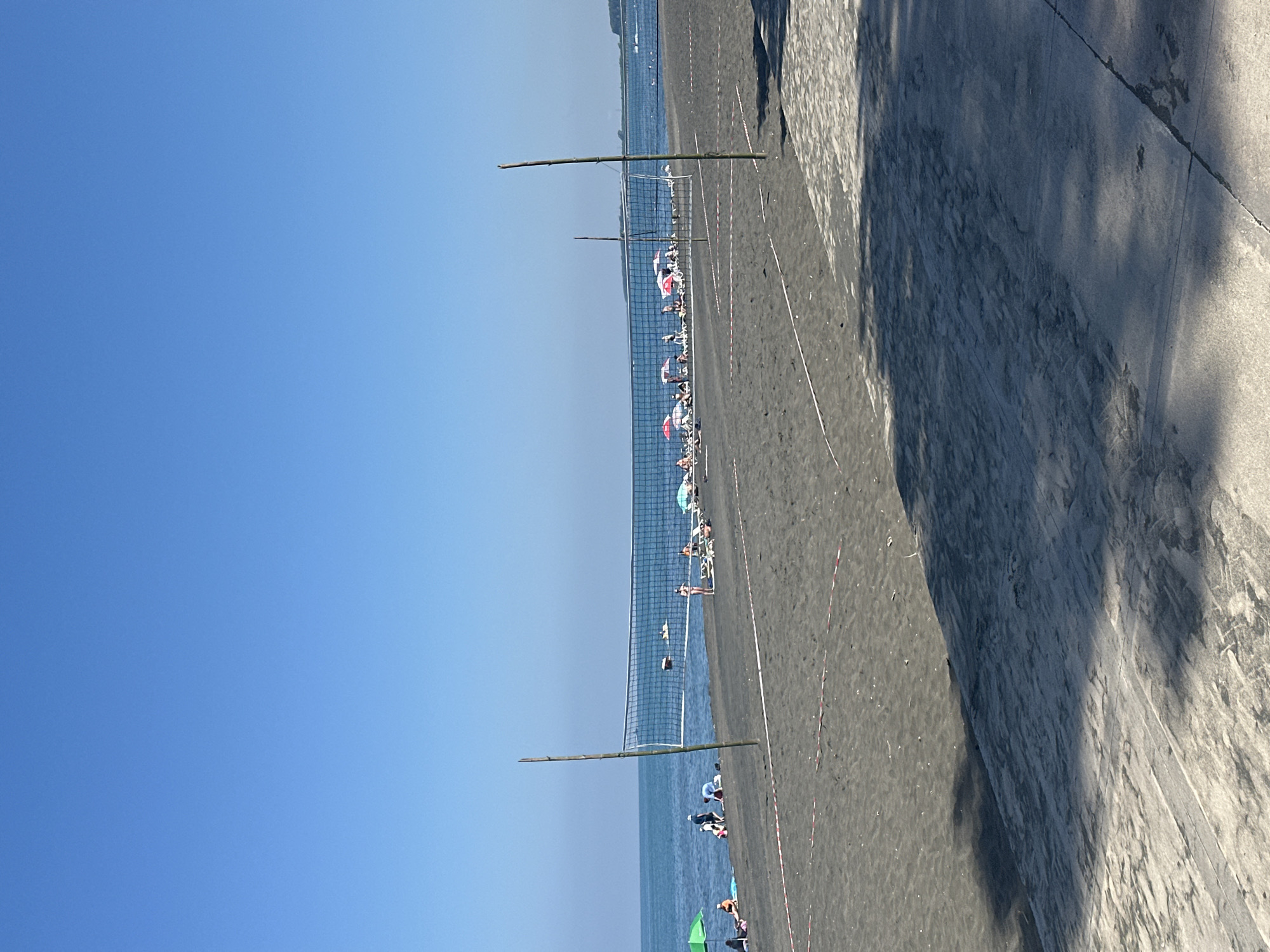 The image depicts a beach scene in Ureki, Georgia, under a clear blue sky. In the foreground, a volleyball net is set up on dark sand, with two bamboo poles supporting it. Red and white caution tape is laid out on the sand around the volleyball court area. Further back, along the shoreline, people are visible relaxing on lounge chairs under colorful umbrellas. The calm sea stretches out to the horizon. The ground in the immediate foreground appears to be a paved walkway or promenade, with shadows cast across it.