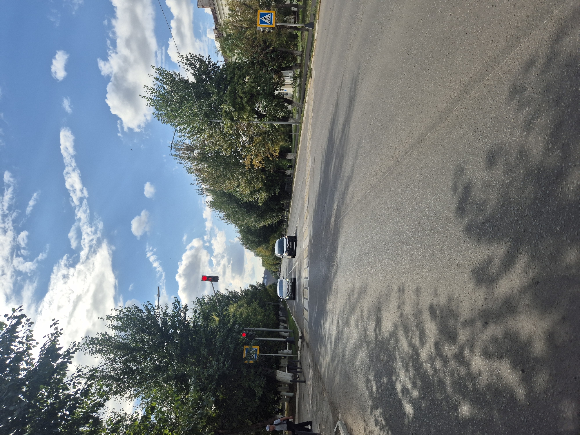 The image depicts a street scene in Kokshetau, Kazakhstan. A paved road runs through the frame, lined with trees on both sides. The sky is blue with scattered white clouds. A traffic light is visible, displaying a red signal. Two cars, a silver sedan and a black van, are stopped at the intersection. A pedestrian crosswalk is marked on the road. To the left, a man in a light-colored cap and dark clothing is walking on the sidewalk. A pedestrian crossing sign is also visible on the left side of the road. On the right side of the road, another pedestrian crossing sign is mounted on a pole. The overall environment suggests a daytime urban setting with moderate traffic.
