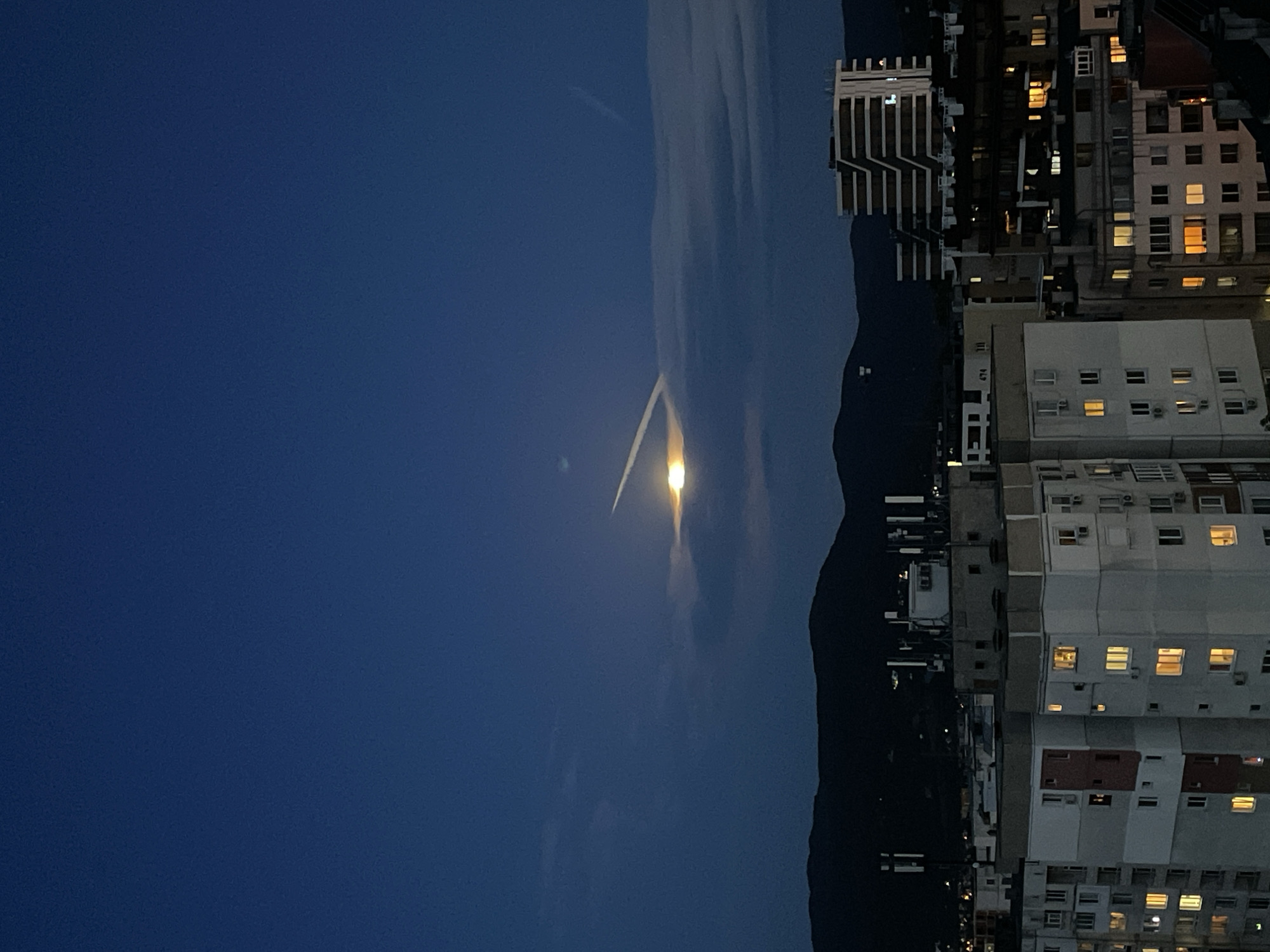 The image shows a night scene in Sofia, Bulgaria. The sky is dark blue, with some light clouds near the center of the image. There is a bright light source, likely the moon, shining through a gap in the clouds, creating a contrail-like effect. To the right, a mountain range forms a dark silhouette against the sky. In the foreground, there are several tall buildings with lit windows, suggesting residential or office spaces. The composition suggests the photograph was taken from an elevated position, perhaps another building. The overall environment appears urban, with a blend of natural elements (mountains, sky) and built structures.