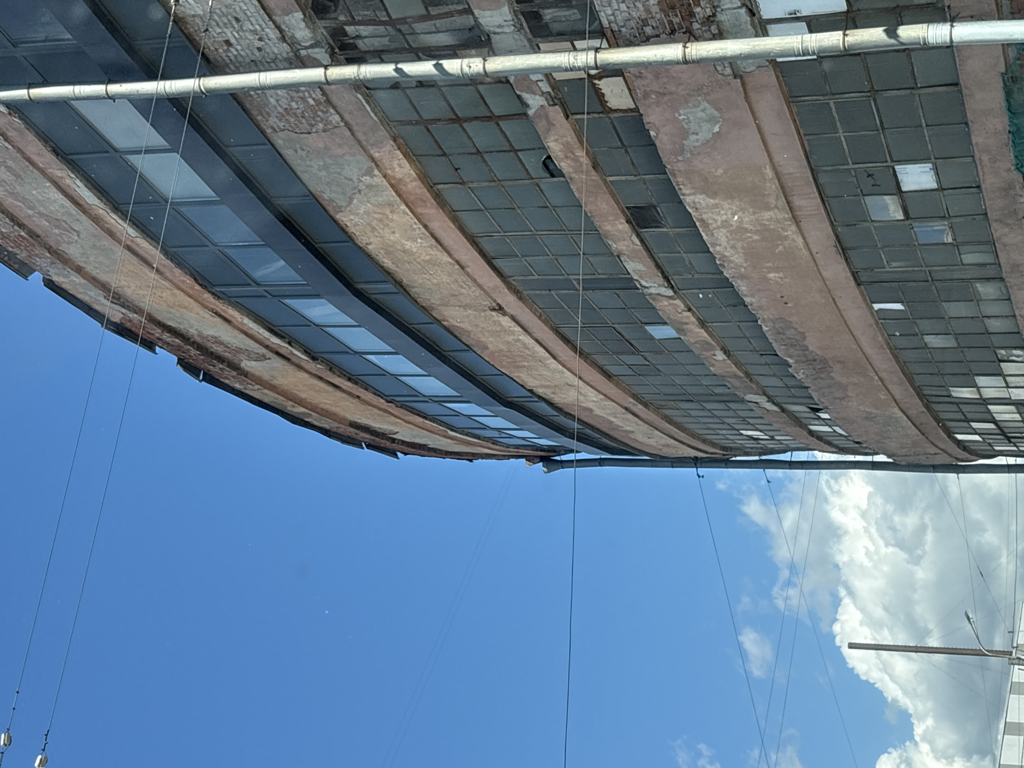 A medium shot captures the curved facade of a multi-story, abandoned industrial building under a bright blue sky with scattered white clouds. The building's exterior is constructed of weathered, pinkish-brown brick, with sections of crumbling plaster revealing the underlying brickwork. Rows of large, multi-paned windows, some filled with dark glass and others broken or missing, run horizontally along each floor. A dark horizontal band runs between two rows of windows. A corrugated metal downspout snakes down the curved side of the building. A silver metal pipe runs vertically along the right side of the building, segmented by horizontal bands. Overhead, thin black wires crisscross the sky. In the lower left corner, a tall, thin pole, possibly a lamppost, is visible against a backdrop of white clouds. The overall impression is one of urban decay and neglect in an industrial district.