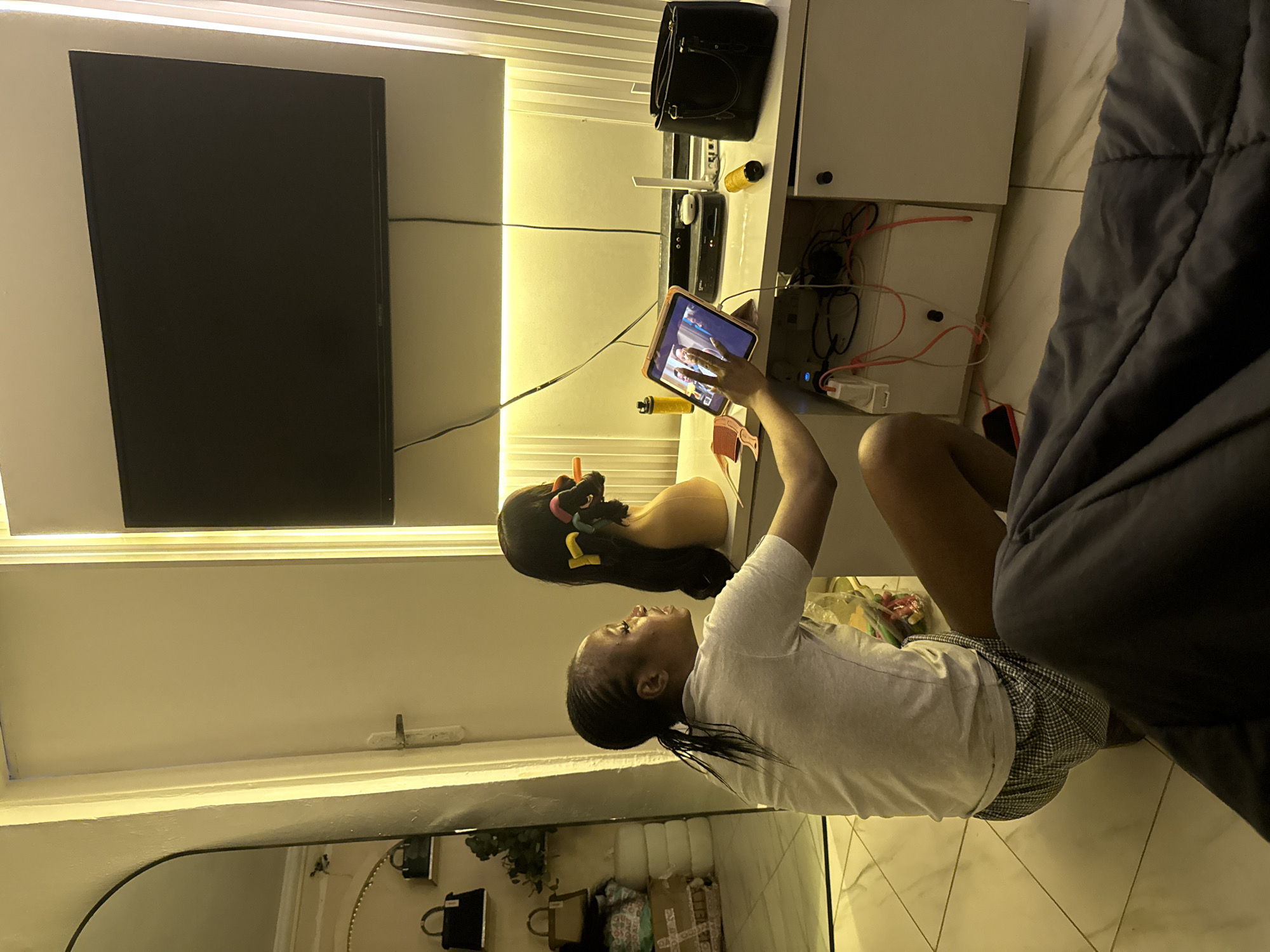 A young Black woman with braided hair and a ponytail, wearing a light-colored t-shirt and checkered shorts, is seated on the floor in a room in Abuja, Nigeria. She is looking at a tablet device that is propped up on a stand on a white cabinet. Her right hand is reaching out towards the tablet, as if interacting with the screen. In the background, a large television is mounted on the wall above the cabinet, and a mannequin head with curlers in its hair is positioned on the cabinet. The room appears to be a bedroom or living area, with tiled flooring and soft lighting. There are various items on the cabinet, including a black handbag, a router, and a water bottle. The overall impression is one of a personal space where the woman is engaged in a relaxed activity.