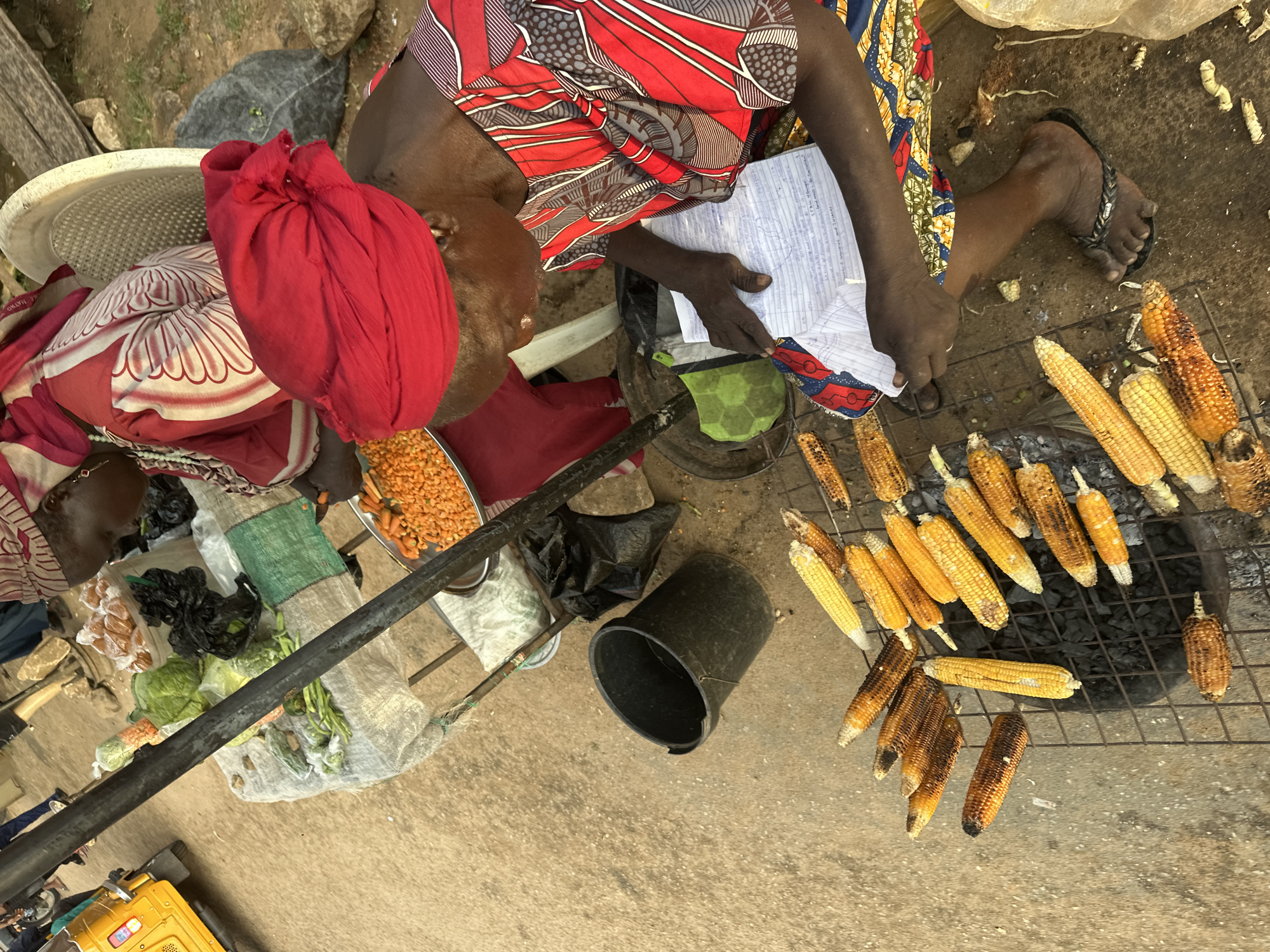 A street vendor in Narayi, Nigeria, grills corn over charcoal on a metal grate. The vendor, a woman with dark skin wearing a red headscarf and a vibrantly patterned dress, is seated on the ground and reviewing notes. In the background, a small yellow motorized rickshaw is visible, as well as a table laden with fresh produce, including cabbage and carrots. The ground is unpaved and dusty. The scene appears to be a typical outdoor market setting.