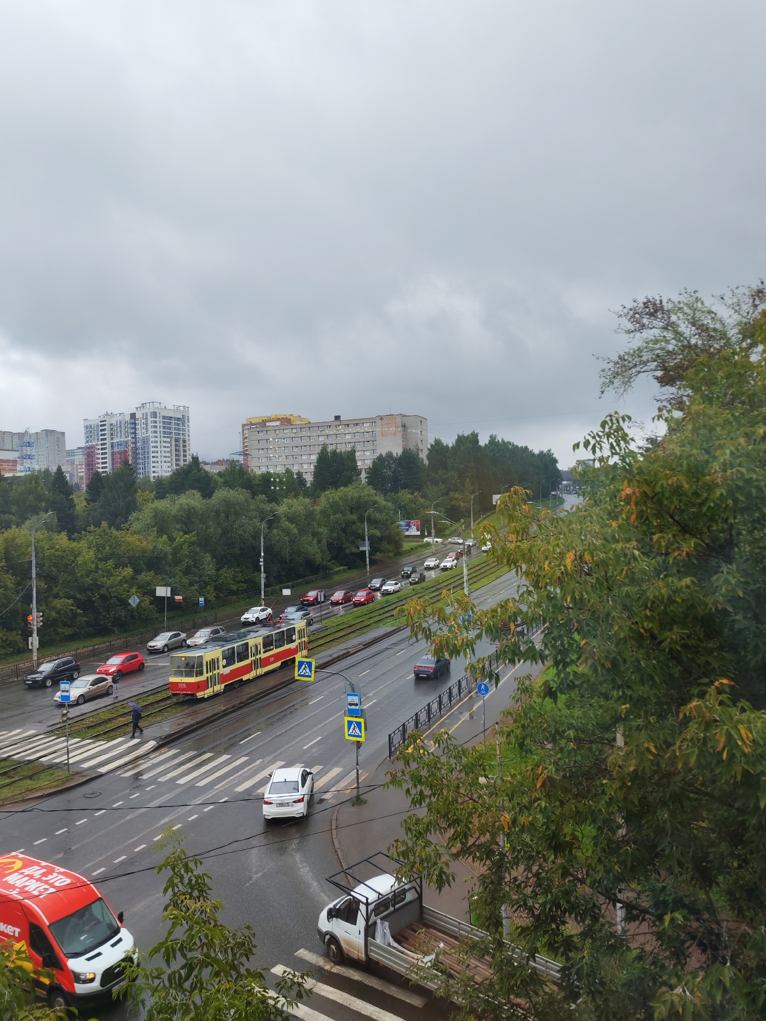 The image depicts an overcast day in Izhevsk, Russia, likely during autumn, with a view of a street scene. In the foreground, a red and yellow tram is on the tracks, with several cars and a white passenger vehicle on the wet asphalt road. There are pedestrian crossings with white stripes and road signs.
Further down the road, traffic is visible, including a red van with white lettering on the side, and a white utility truck with an open back. Trees with green and some yellowing leaves line the sides of the road, and behind them, several apartment buildings of varying heights and architectural styles are visible, including modern high-rises and older, block-style buildings. The sky is heavily clouded and gray. A person with an umbrella is walking across a pedestrian crossing. The wet condition of the road suggests it has recently rained or is raining. The scene appears to be a busy urban intersection.
