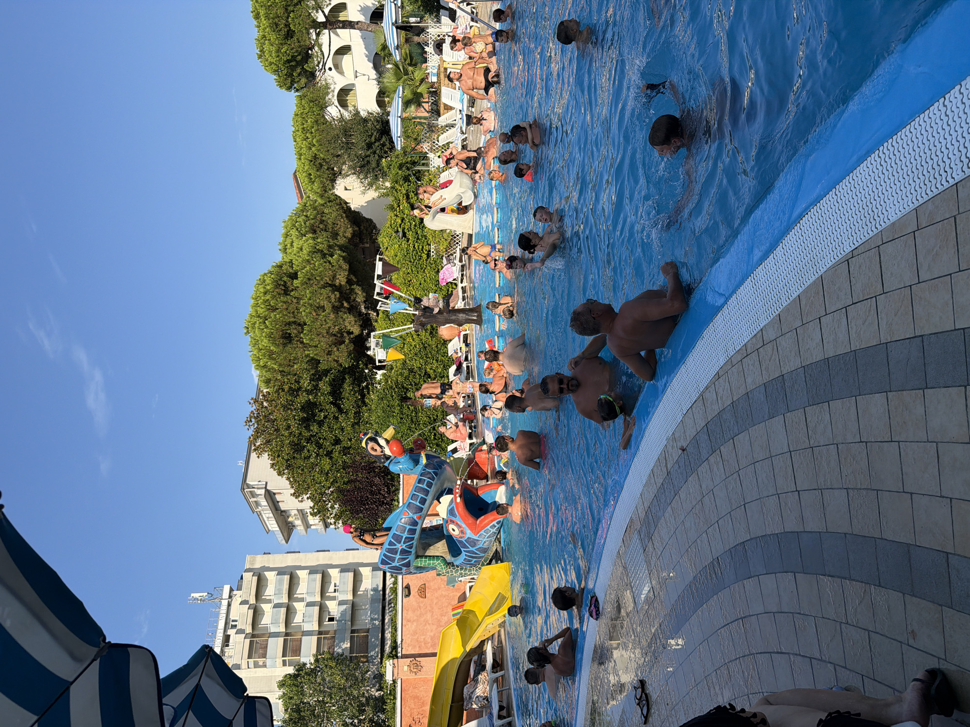 The image depicts a crowded outdoor swimming pool on a sunny day. The pool is filled with a diverse group of people, including adults and children, engaged in various water activities. Some individuals are swimming, while others are relaxing or playing in the water. There's a large, colorful water slide in the background, a popular attraction. Umbrellas and lounge chairs are visible around the pool, suggesting a resort or hotel setting. In the background, buildings and lush green trees are present, indicating an urban or semi-urban environment. The scene is likely from a tourist area, possibly a hotel complex or a public water park. Given the context provided, this scene is located in the city of Gatteo, Italy.