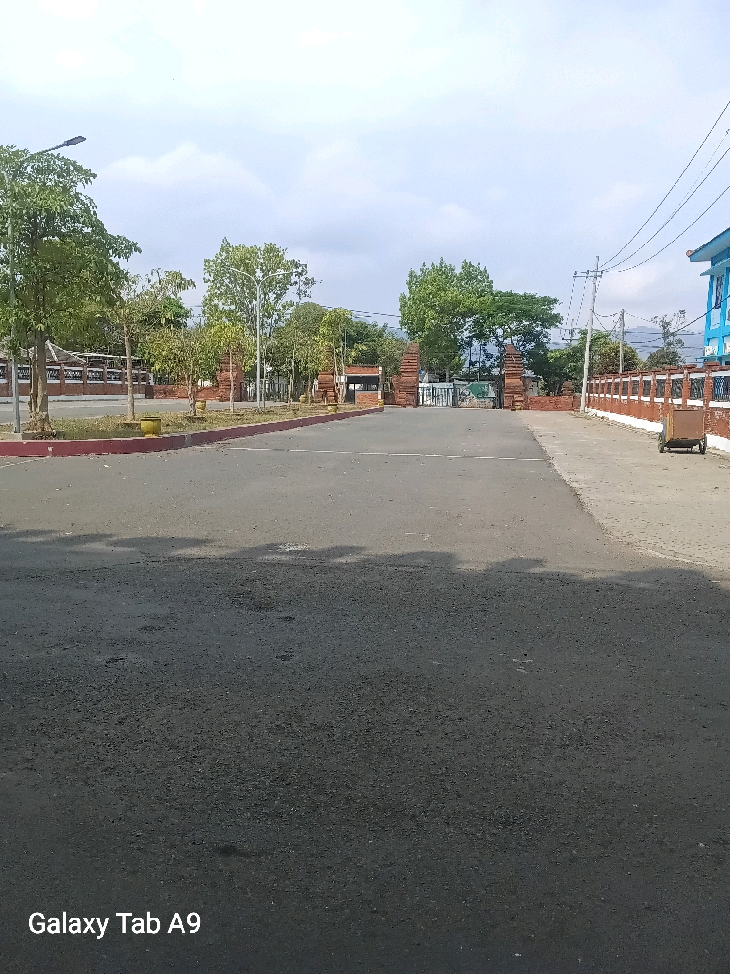 The image depicts an outdoor scene in the city of Trenggalek, Indonesia. A paved road dominates the foreground, leading towards a gateway with brick pillars on either side. Beyond the gateway, there are trees and buildings. To the right of the road, a brick wall with decorative metal fencing runs along the perimeter. A small, wheeled cart is visible near the wall. The sky is cloudy. The overall impression is of an entrance area to a facility or compound.