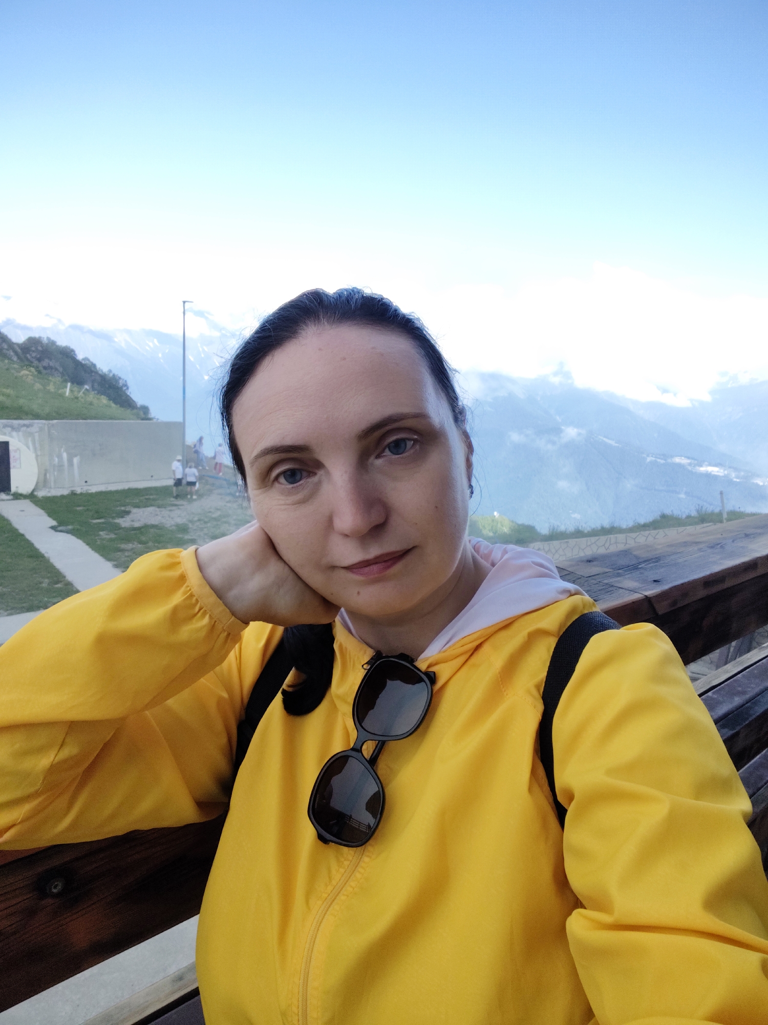 A woman with dark hair pulled back is taking a selfie outdoors. She is resting her head on her hand, with a slight smile on her face. She is wearing a bright yellow zippered jacket with sunglasses hanging from the collar. The background shows a mountainous landscape with a clear blue sky. There are some clouds in the distance, obscuring parts of the mountains. A wooden bench or railing is visible in the foreground. Further in the background, there are some people walking and a pathway leading towards a concrete structure. The lighting suggests it is daytime, likely late afternoon or early evening given the soft light on the mountains. The overall mood is relaxed and pleasant, possibly indicative of a tourist enjoying a scenic view. There is no visible text in the image. The location appears to be a scenic viewpoint in a mountainous area, consistent with Esto-Sadok, Russia.