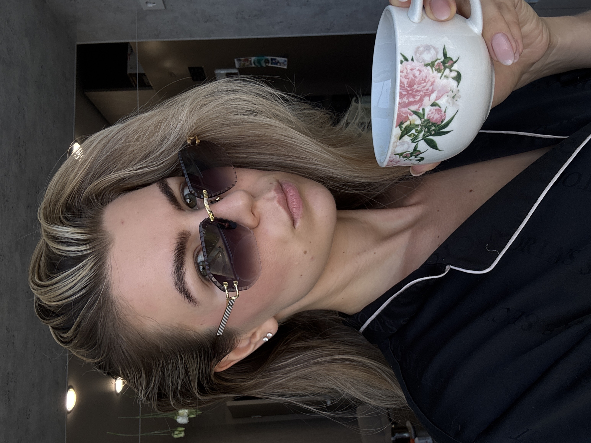 A woman wearing sunglasses and a black pajama top with white trim is holding a white floral-patterned mug in her right hand. She is positioned indoors against a reflective surface that shows parts of the room. The city and specific location are not discernible from the image.