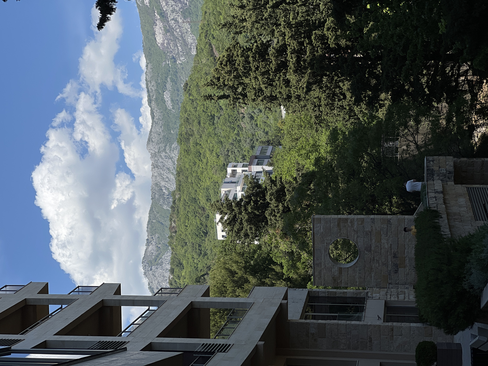 This landscape image captures a serene view of a mountainous region, likely Boreti, Montenegro, under a bright blue sky adorned with scattered white, puffy clouds. The foreground features prominent architectural structures. To the left, a modern building displays a distinctive concrete facade with a grid of angular, open frames, suggesting balconies or architectural details. To its right, a stone-clad building stands out with a large, perfectly circular opening in its upper section. Lush green hedges line the base of this stone structure.
Two individuals are subtly visible in the image. One person, wearing a yellow shirt, is partially obscured by the hedge near the stone building. Further to the right and slightly elevated, another person in a white shirt is standing on a balcony or platform, mostly hidden by dense green foliage.
The mid-ground is dominated by a rich expanse of diverse green trees, including tall conifers, which densely cover the mountainous terrain. Nestled among these trees, several smaller, white-colored buildings with flat roofs can be seen, blending into the natural environment. In the background, majestic gray mountains rise, their rugged, rocky peaks contrasting with the vibrant green slopes below.
The scene appears to be set on a clear, sunny day, likely during the morning or early afternoon, given the bright, even lighting and the shadows cast by the trees and buildings. The overall impression is one of peaceful coexistence between modern architecture and a beautiful natural landscape. There is no visible text in the image.