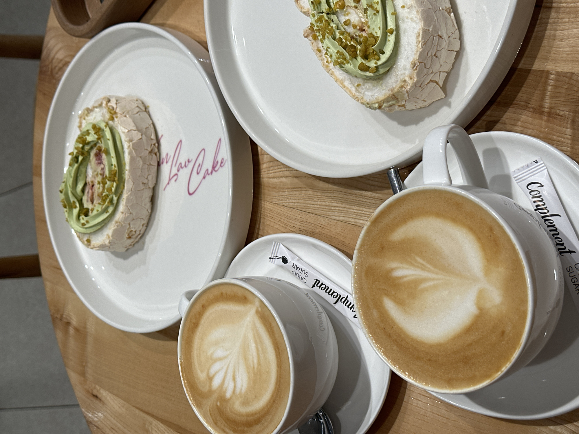 An eye-level shot features two plates of green-frosted desserts, alongside two cups of coffee with latte art, and sugar packets, set against a wooden table. The desserts are rolled and garnished with green frosting and chopped nuts. The plate on the left has pink cursive text that says "In Law Cake." Both cups are filled with coffee, adorned with a latte art design. Each cup is placed on a saucer accompanied by a single-serving sugar packet from "Complement." The wooden table provides a warm-toned background. It’s difficult to determine a specific location in Rostov-on-Don, Russia, based on the image, as it is an interior shot of a cafe or similar establishment. There are no people visible in the image, nor actions or interactions taking place. The environment is a clean, well-lit cafe.