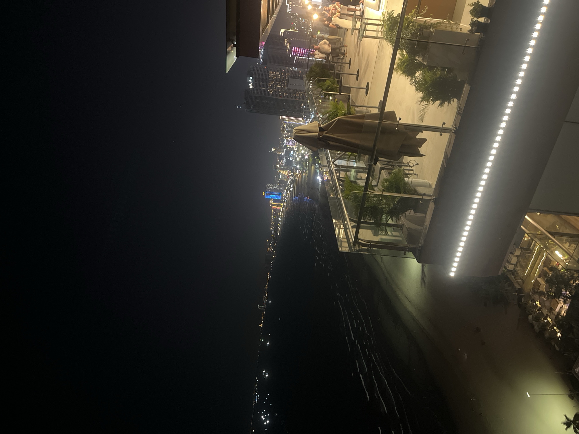 This media file depicts a nighttime scene at a rooftop establishment in Đà Nẵng, Vietnam, overlooking a city skyline and a body of water. The camera angle is tilted, giving a sense of looking down and out towards the illuminated cityscape.
In the foreground, there are tables and chairs, some with umbrellas, suggesting an outdoor dining or lounge area. People are visible in the background, gathered at tables and socializing, their forms illuminated by the ambient light from the city and the establishment's lighting. Plants are strategically placed around the area, adding to the ambiance.
The main focus is the vibrant cityscape at night. Numerous skyscrapers are lit up, creating a dazzling display of lights against the dark sky. The reflections of these lights can be seen on the surface of the water, which stretches out towards the city. A prominent blue-lit structure stands out in the distance. The overall mood is lively and sophisticated, indicative of a popular urban nightlife spot. The time of day is clearly night, and the weather appears clear, allowing for an unobstructed view of the city. The presence of a beach or coastline is suggested by the dark, rippling expanse in the lower portion of the image, which could be the sea or a wide river. There is no visible text in the media file.