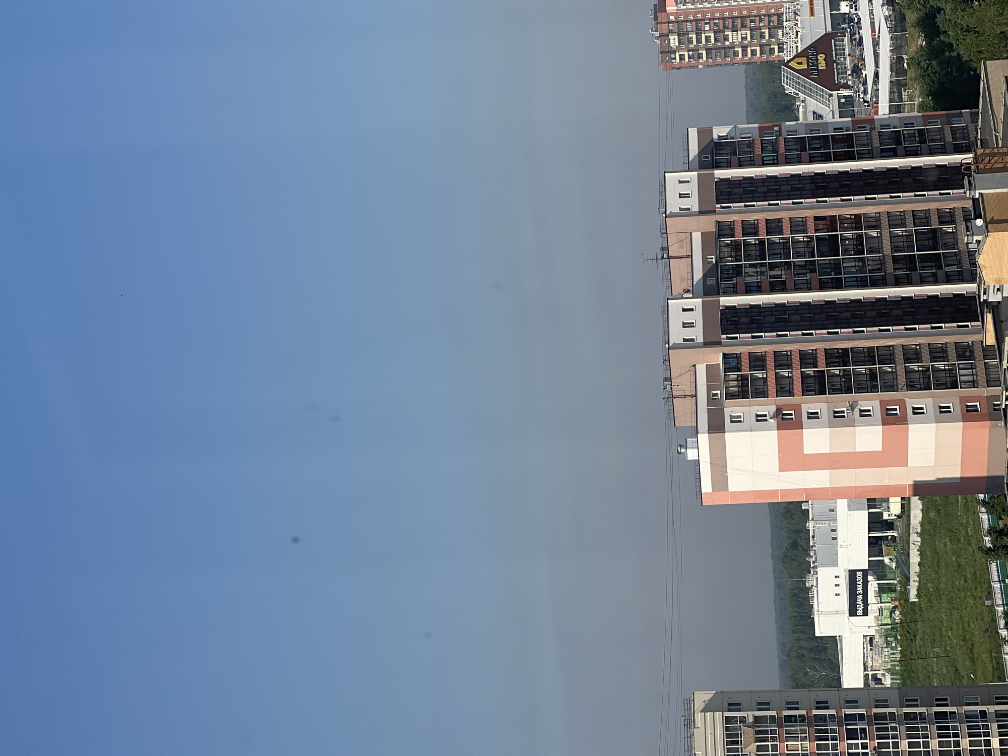 This is a distant aerial view of a residential area in Novosibirsk, Russia, on a clear, sunny day with a blue sky. The foreground is dominated by the tops of several high-rise apartment buildings. One building on the left has a white and pink exterior with many windows. Another larger building in the center is also tall with a white, pink, and brown facade and numerous windows. To the right of this, a taller, more densely windowed building with a red-brown exterior is visible. In the middle ground, there is a long, white building with a dark sign that reads "ВЫДАЧА ЗАКАЗОВ" (Order Pickup). Beyond this building, a treeline of green forest stretches across the horizon. Further to the right, there is a shopping center with a distinctive A-frame roof and a parking lot. The scene is devoid of any visible people or animals. The environment appears urban, with modern architecture and infrastructure.