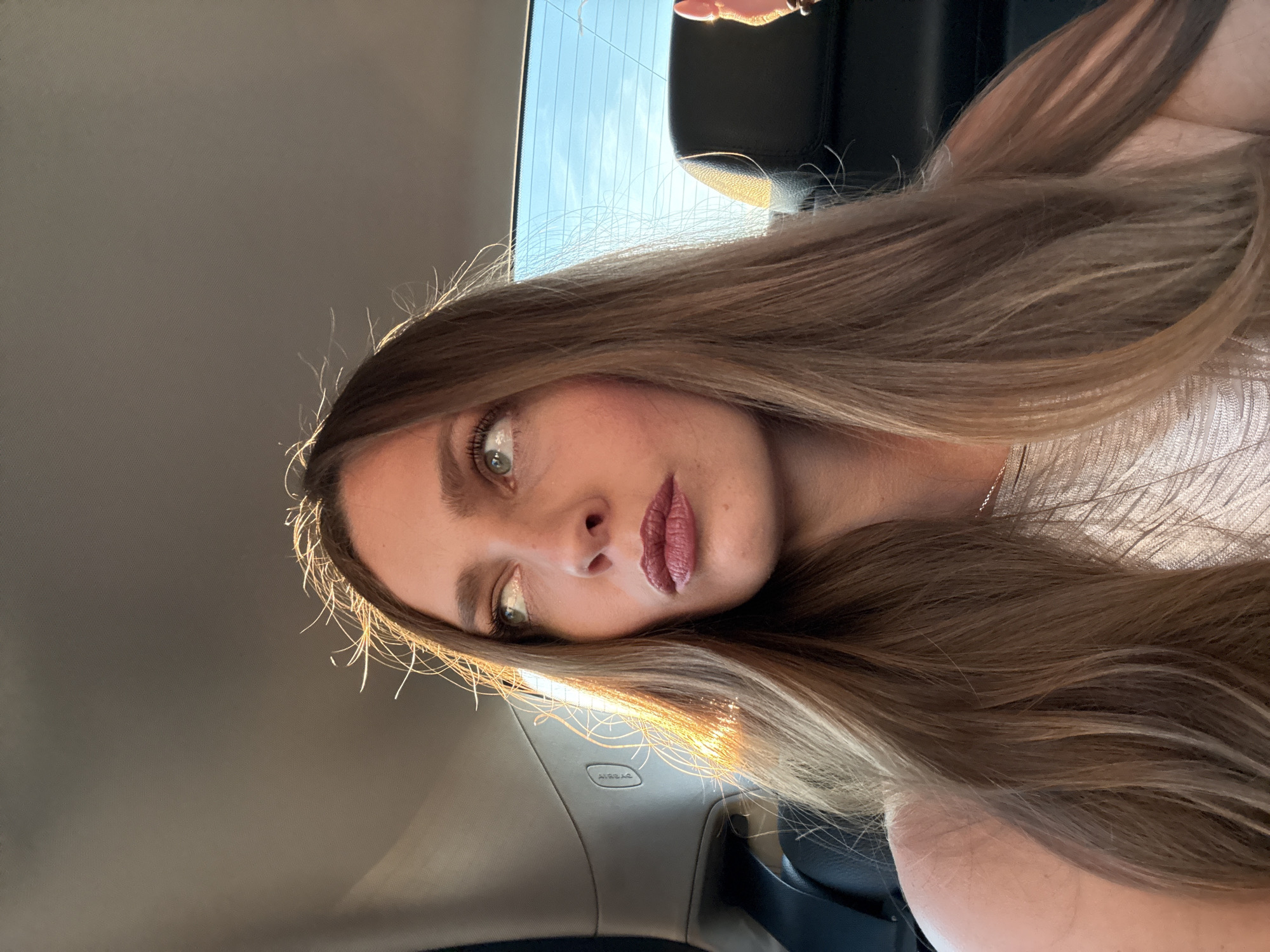 A young adult woman is seated in the rear passenger compartment of a vehicle, in Moscow, Russia. Her long, light-brown hair, exhibiting flyaways highlighted by strong golden hour sunlight entering from the left, frames her face. She wears a light-colored, possibly textured garment with bare shoulders and a thin chain necklace, along with dark berry-toned lipstick. Her eyes, light in color, are directed upwards and to the left, gazing out of frame with an observant or pensive expression. The car interior features black leather seats, a light gray headliner, and a visible "AIRBAG" label on the B-pillar. Through the rear windshield, a clear blue sky with faint horizontal lines (possibly power lines or distant contrails) is visible.