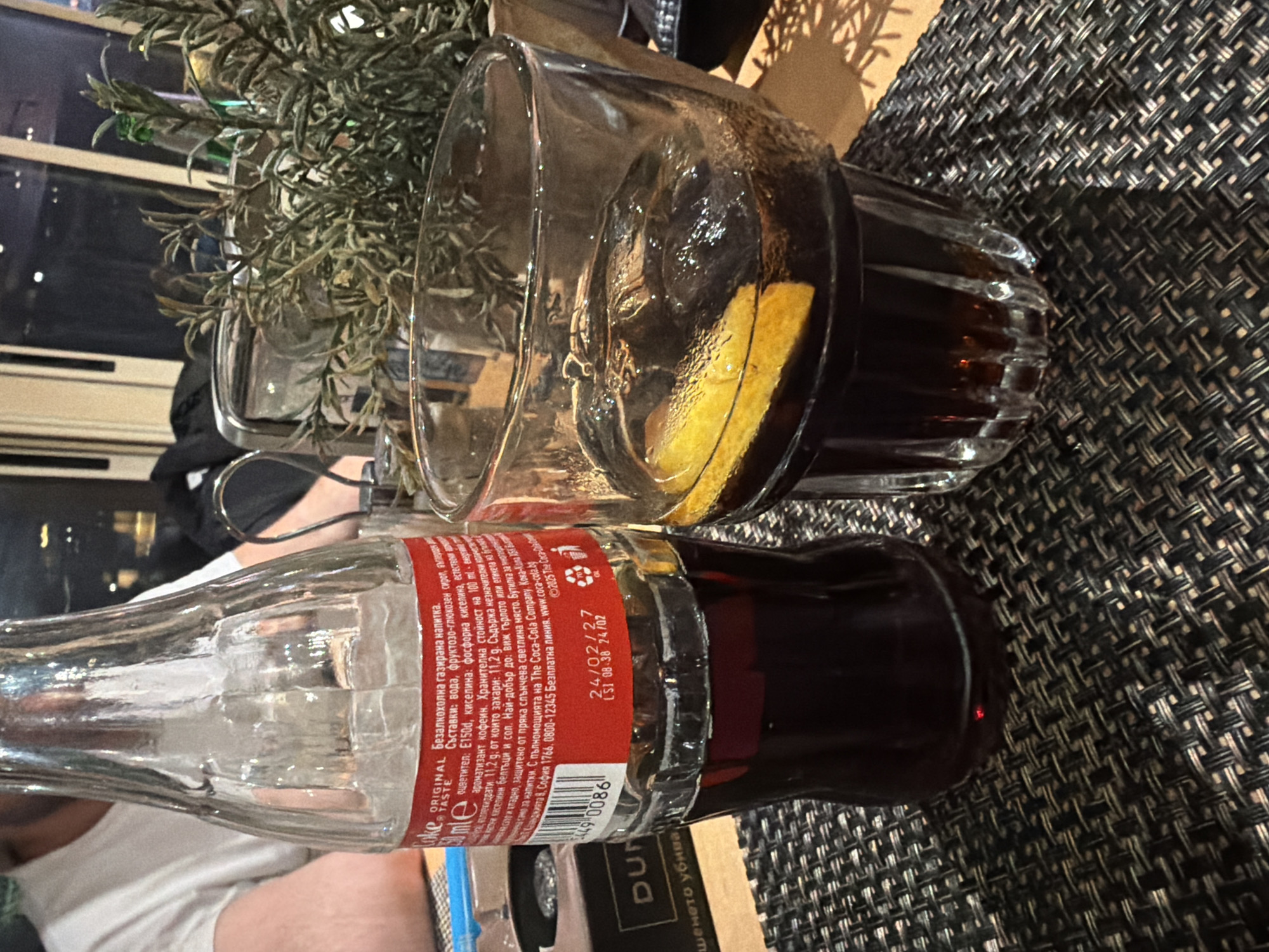 The image depicts a glass of Coca-Cola with ice and a lemon slice, next to an open Coca-Cola bottle. The items are placed on a patterned placemat. In the background, out-of-focus, there is greenery. The text on the Coca-Cola bottle is in Bulgarian, and it includes a best-by date of 24/02/27. There is no specific information provided to conclude this is in the city of Plovdiv, Bulgaria.