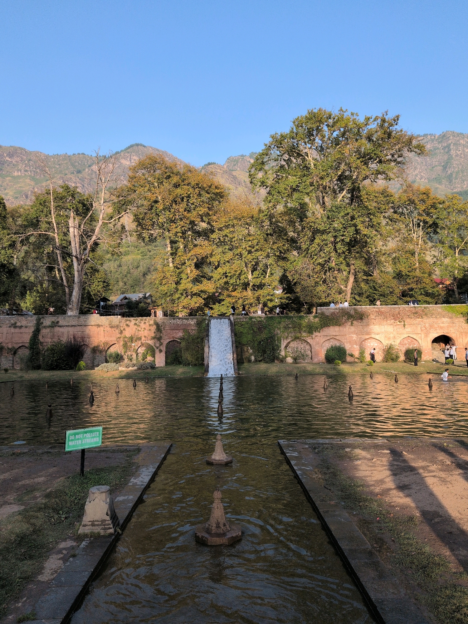 A wide, eye-level shot captures a tranquil scene in the Shalimar Bagh Mughal Gardens in Srinagar, India. The pristine blue sky provides a clear backdrop to the lush greenery of the surrounding landscape and the architectural features of the garden. In the foreground, a reflective pool of water mirrors the golden hues of the late afternoon sun. Several small, dark fountains dot the surface of the water, adding a subtle ripple to the stillness.
A paved pathway extends into the water from the bottom edge of the frame, with a narrow stone embankment leading the eye towards a more expansive section of the garden. To the left, a green sign with white lettering reads "DO NOT POLLUTE WATER STREAMS."
The middle ground is dominated by a raised, brick-lined terrace with arched openings, characteristic of Mughal architecture. A cascading waterfall tumbles down from the terrace into the pool, creating a dynamic contrast to the otherwise serene environment. On the terrace, several people are visible, some walking and others gathered in small groups, enjoying the picturesque setting.
In the background, a dense canopy of mature trees, with foliage in shades of green and golden yellow, suggests the autumn season. Beyond the trees, the rugged slopes of distant mountains rise up, their contours softened by the hazy sunlight. The overall atmosphere is one of peacefulness and natural beauty, enhanced by the harmonious integration of water, greenery, and historical architecture.