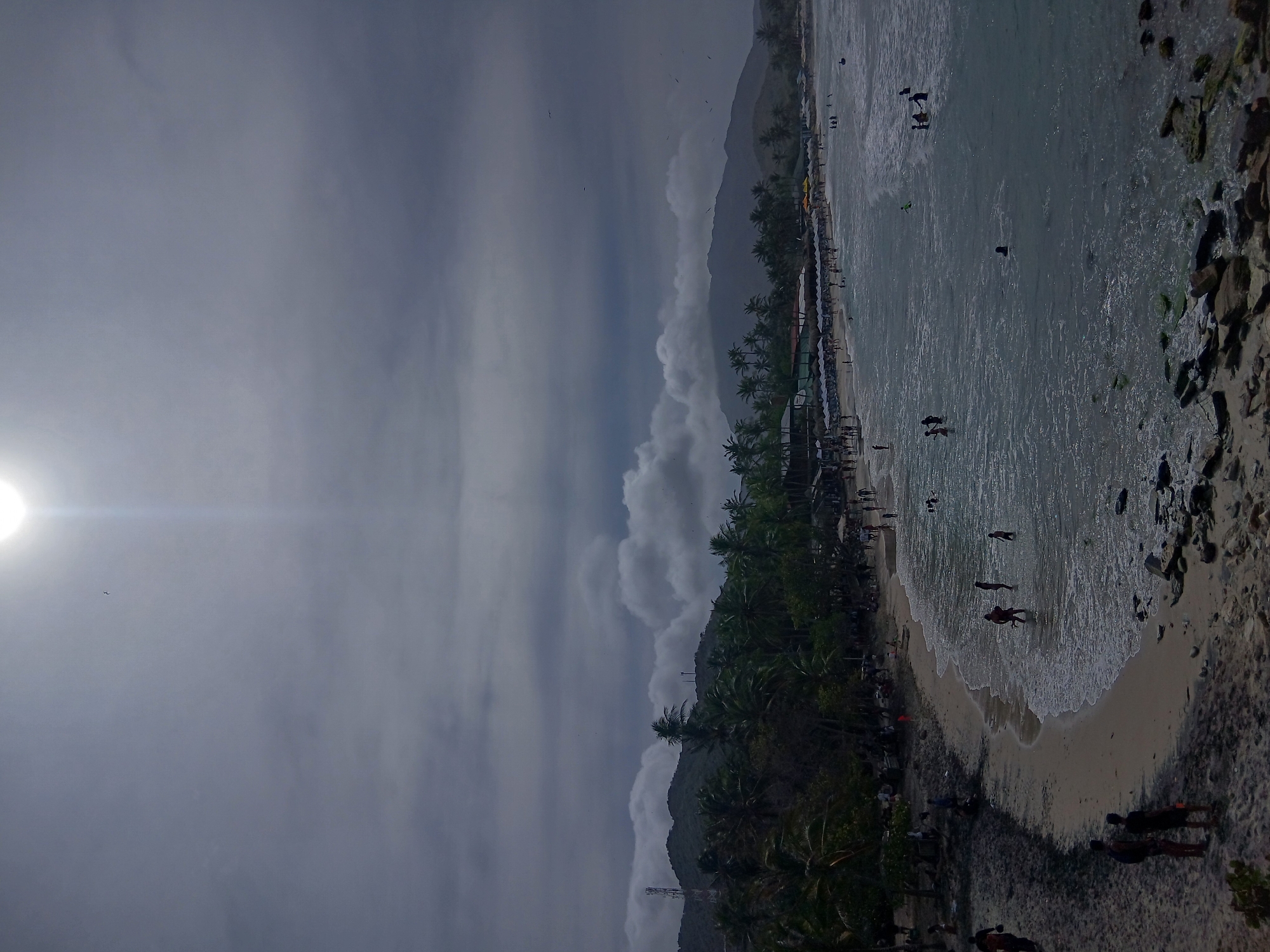 The image captures a view of Playa Parguito in Venezuela. A beach lined with palm trees meets the ocean, where people are swimming and wading in the water. The beach has a sandy area where people are standing or walking, and there are also rocks on the shore. On the left, the sun is obscured by clouds, with its light streaking across the sky. Mountains are visible in the background, partially obscured by clouds. The sky is overcast.